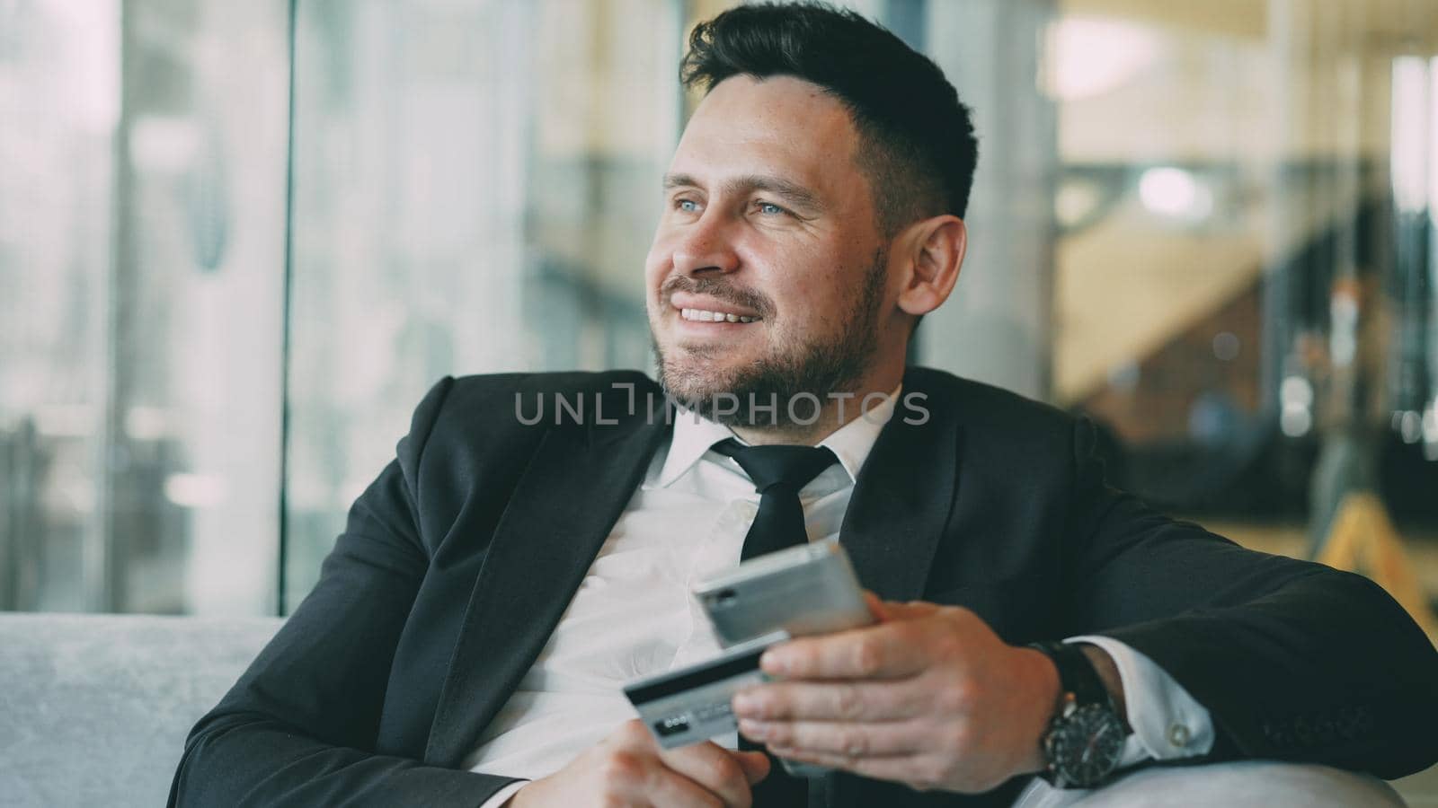Cheerful Caucasian businessman in suit using online banking holding credit card and smartphone in his hands in modern cafe indoors during lunch break