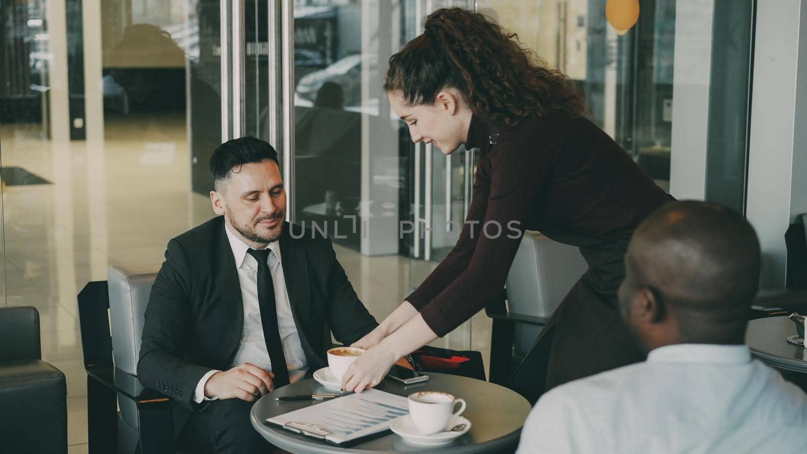 Cheerful Caucasian businessman in formal clothes smiling and discussing business profit with his African American colleague in cafe. Young waitress bringing cup of coffee. by silverkblack