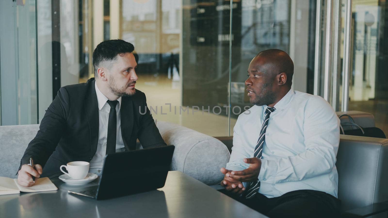 Two confident multiracial businessmen discussing about partnership during meeting in modern glassy cafe