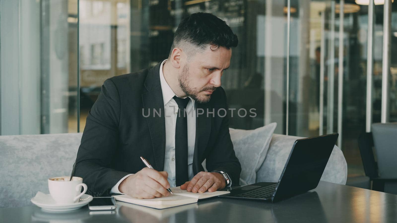 Smart Caucasian businessman using laptop computer and writing down information in his notepad in glassy cafe having coffee cup on his table. A cup of coffee is on his table.