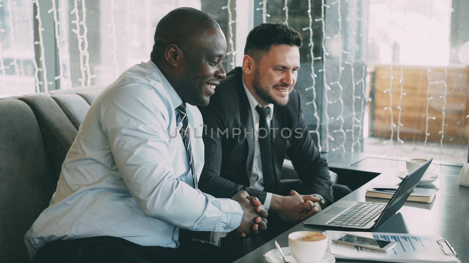 Cheerful African American businessman in formal clothes discussing business project with his caucasian colleague on his laptop in cafe by silverkblack