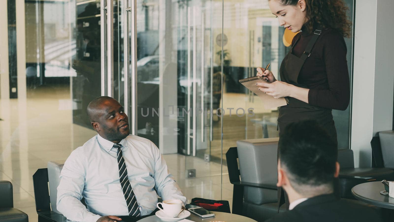 African American businessman smiling, drinking coffee and discussing startup with his Caucasian partner in classy cafe. Young waitress taking order by silverkblack