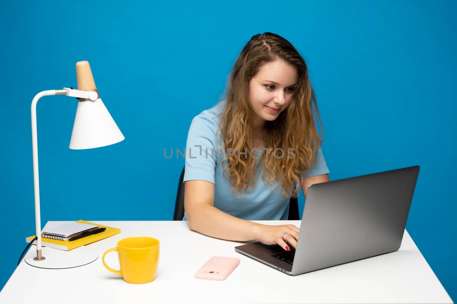 Young curly woman in a blue t-shirt studying from studio using laptop computer over a blue background. Serious beautiful young woman typing on laptop in a bright modern office