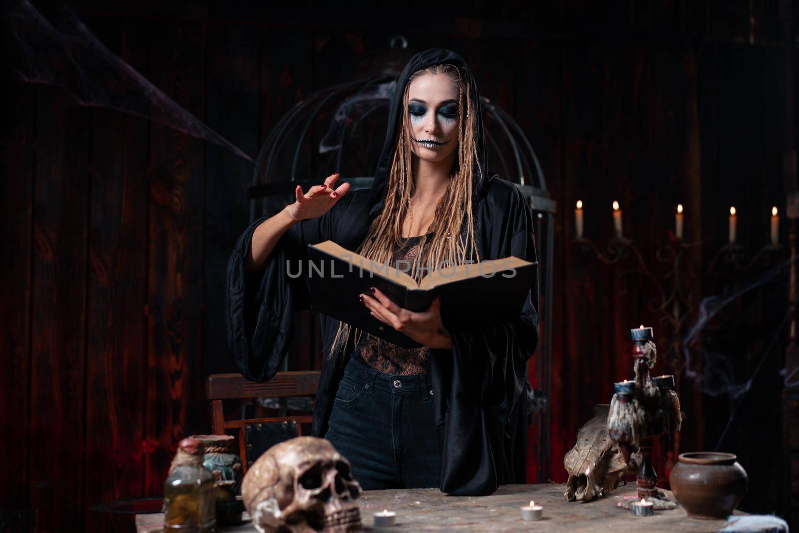 Halloween concept. Witch dressed black hood standing dark dungeon room use magic book for ritual devil calling. Female necromancer wizard gothic interior with skull, cage, spider web