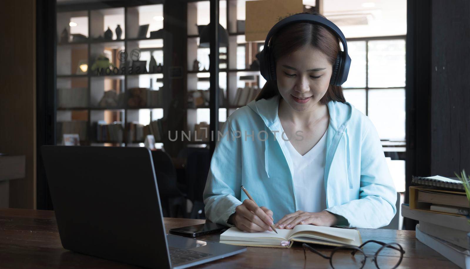 Smiling Asian young female using headset looking at laptop screen listen and learning online courses. Happy chinese business woman with headphones video call for customer service by wichayada