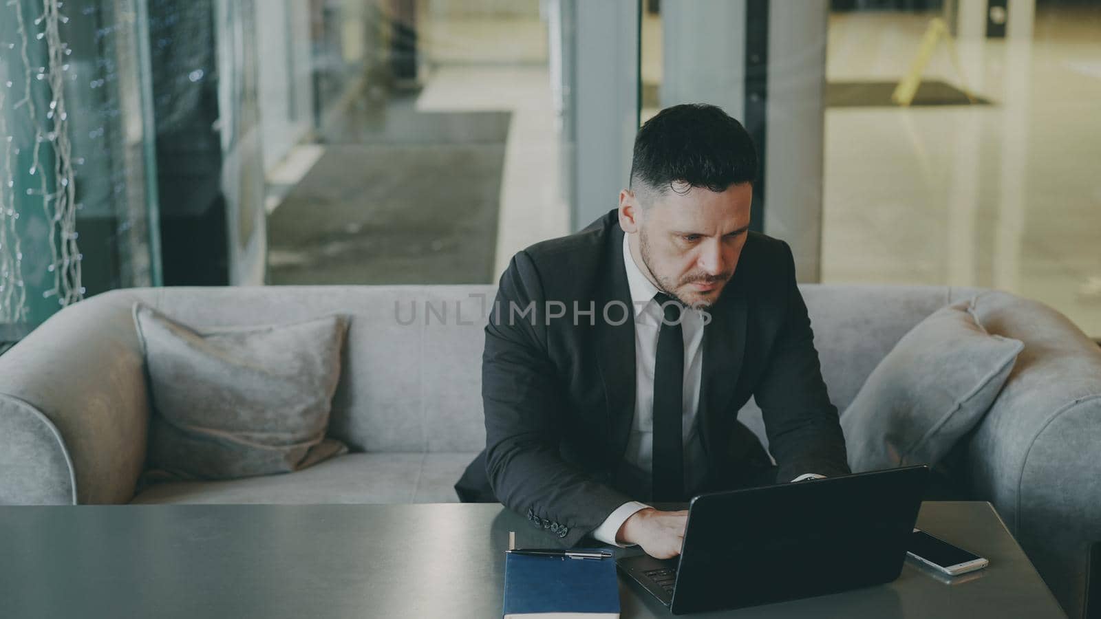 Respected Caucasian businessman in black business suit sitting and working concentrated on his laptop computer in modern cafe indoors