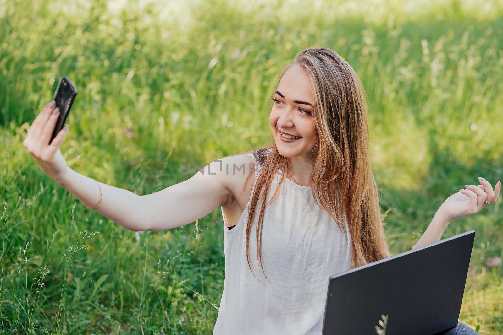 girl sits outdoors and works at a laptop. makes a video call on the phone. freelance. selfeducation. the concept of remote learning and outdoor work. by Anyatachka