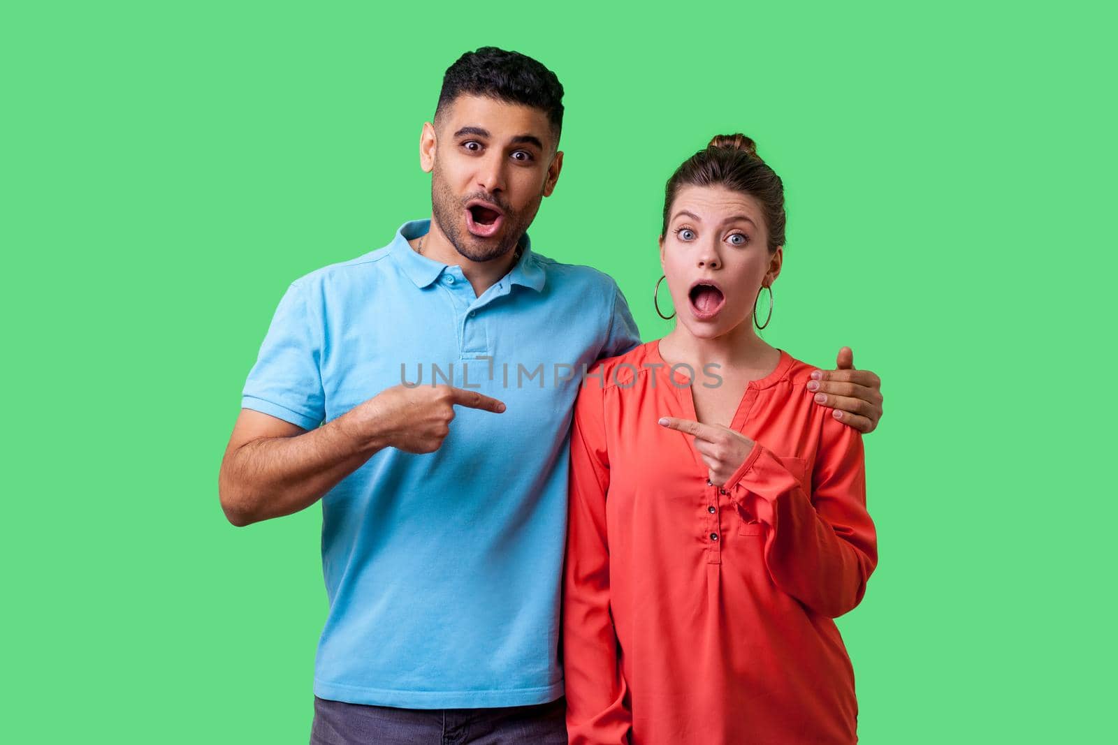 Portrait of amazed attractive young couple in casual wear standing with opened mouths, hugging as friends, pointing fingers and choosing each other. isolated on green background, indoor studio shot