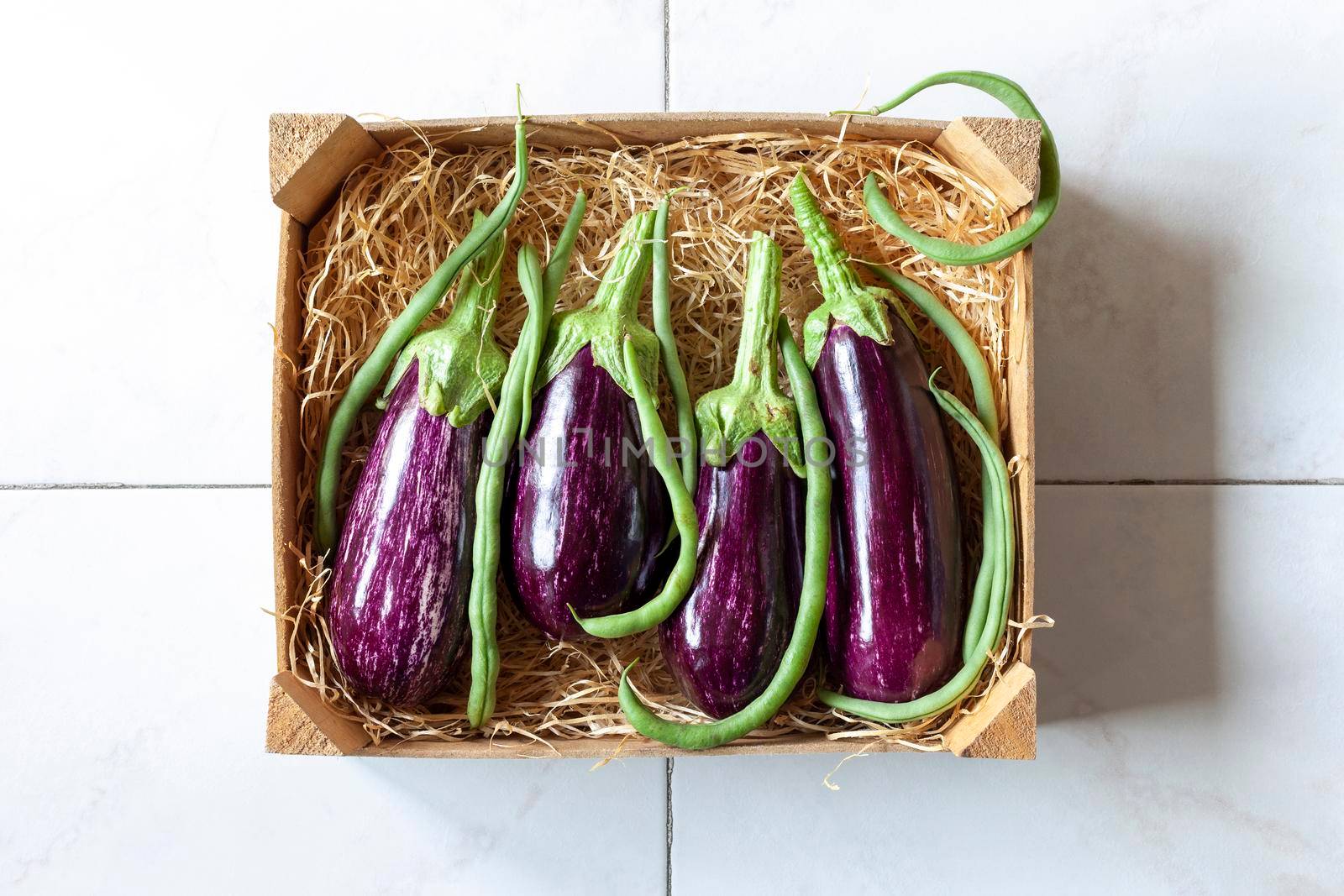 eggplants and green beans in a vegetable wooden case on tiles background, idea of harvest time, top view