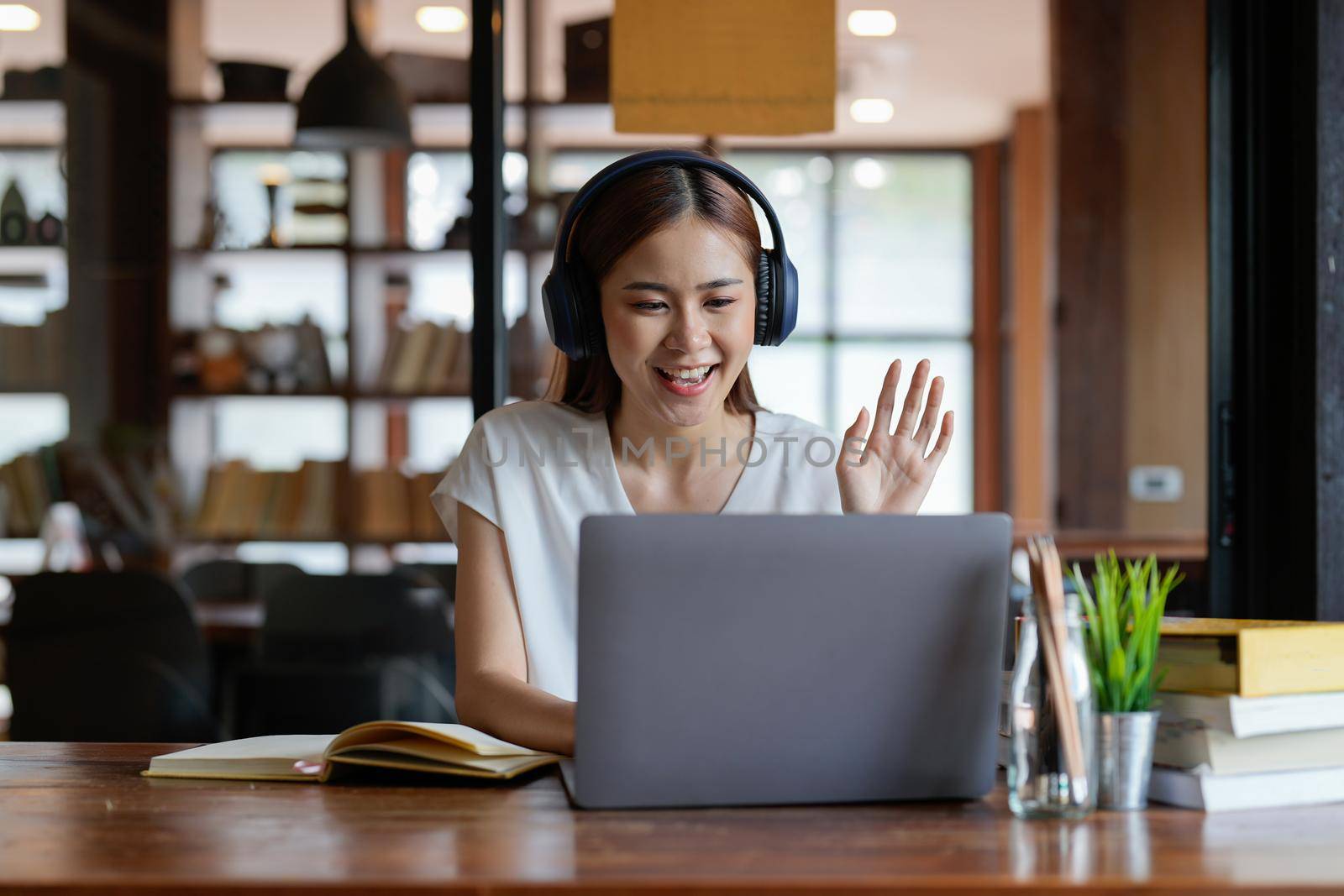 Online education. A female student with headphones learning online at home, she waving hand to other students or colleagues on computer monitor in video conference and smiling. by nateemee