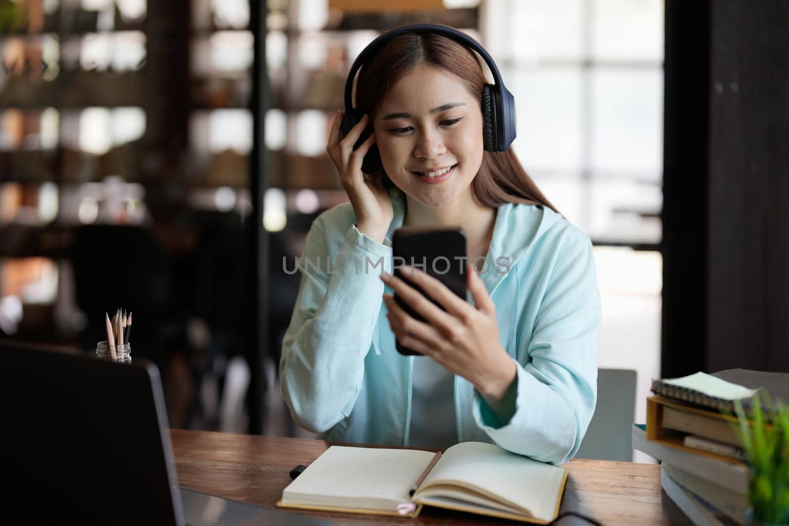 Happy asian student girl in wireless headphones using learning app on mobile phones, watching webinar on smartphone, listening to audio course, studying at open book, copy book at home by nateemee