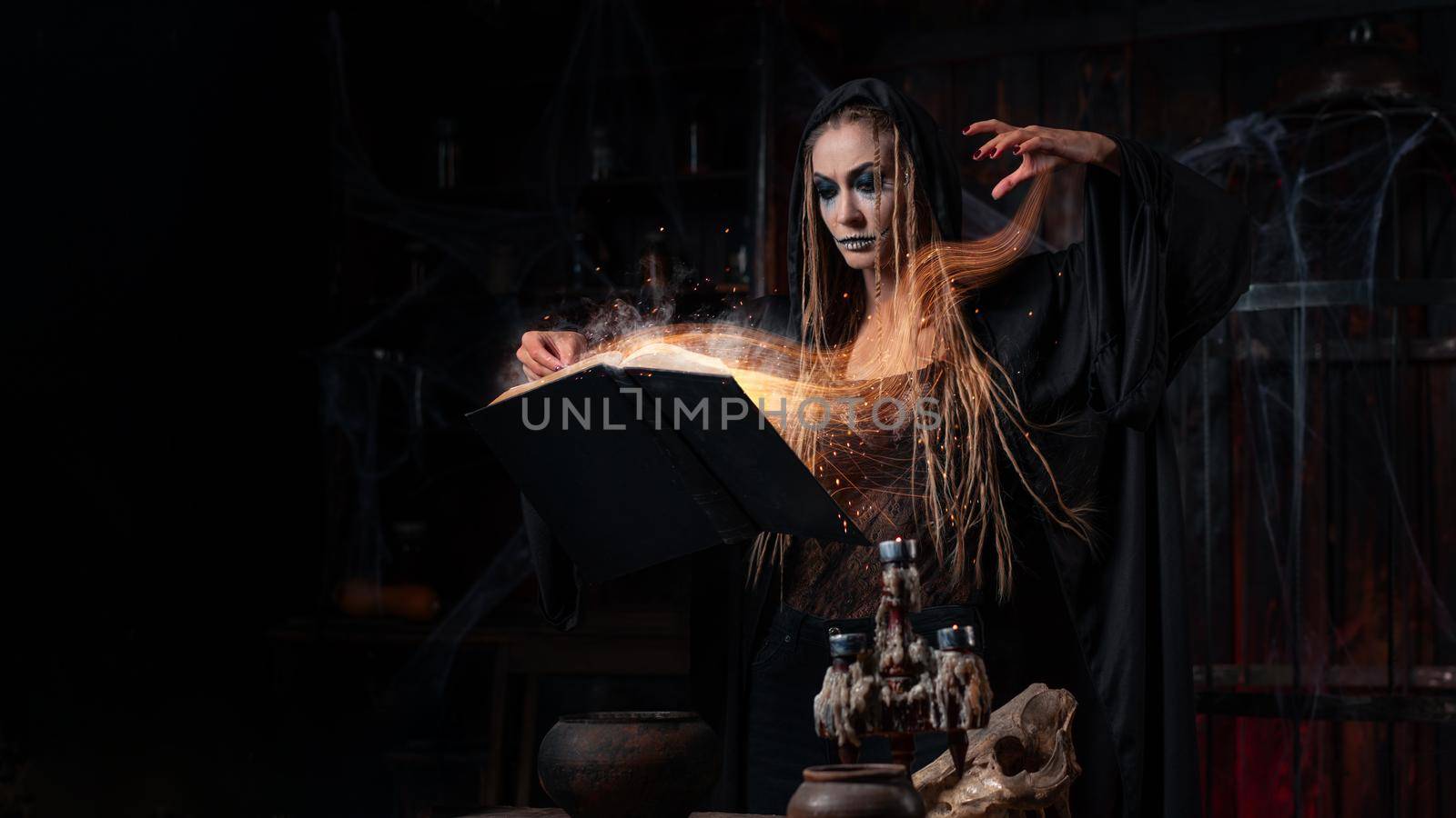 Halloween concept. Witch dressed black hood with dreadlocks standing dark dungeon room use magic book for conjuring magic spell. Female necromancer wizard gothic interior sparkles soul effect