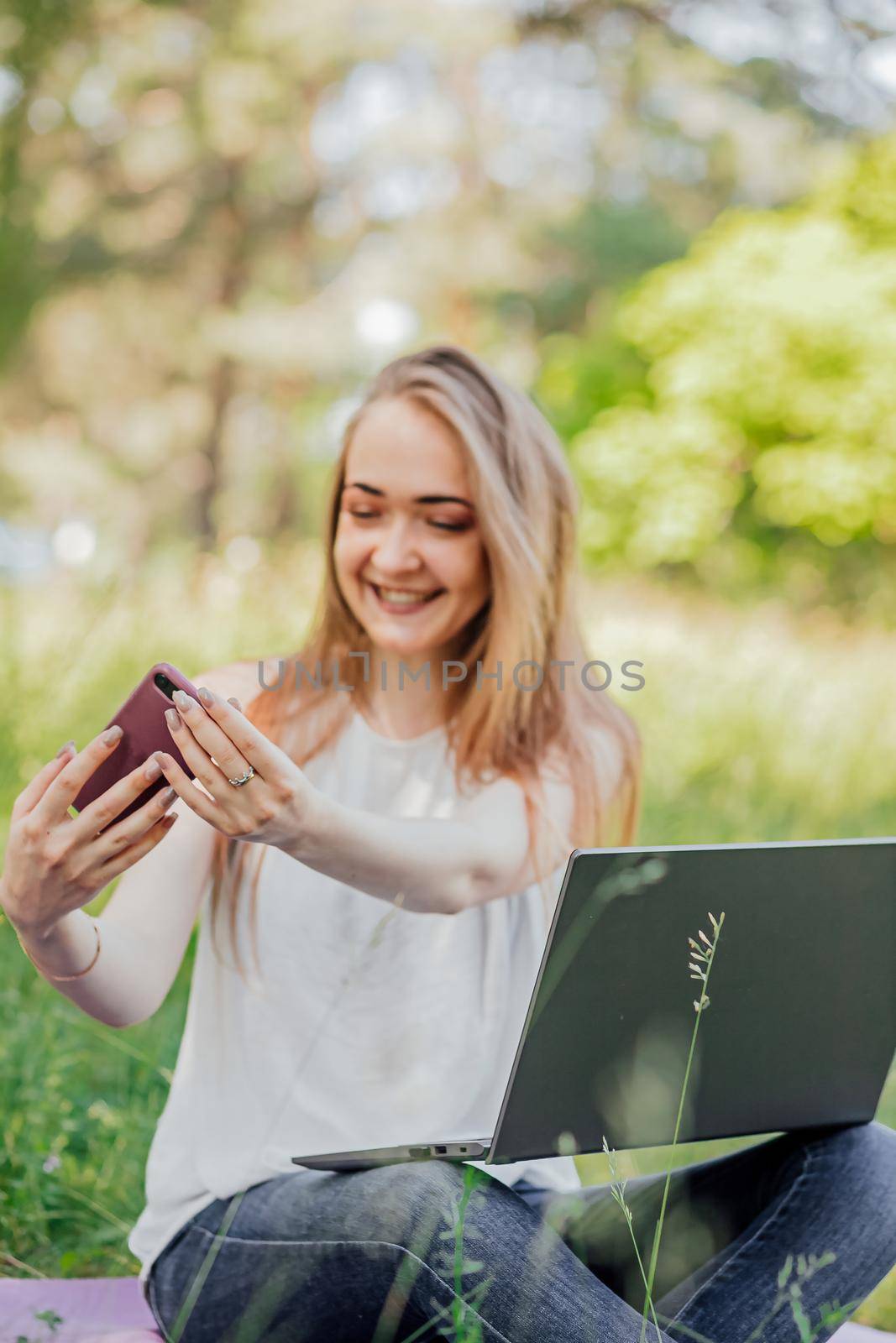 girl sits outdoors and works at a laptop. makes a video call on the phone. freelance. selfeducation. the concept of remote learning and outdoor work. by Anyatachka