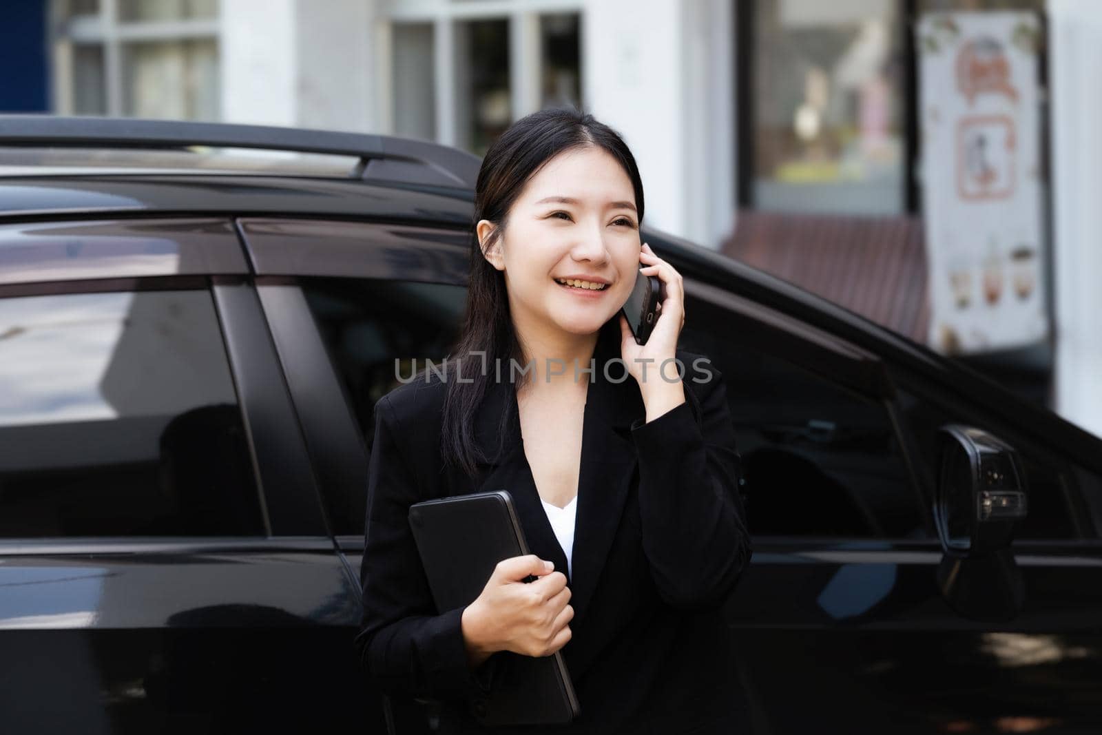 Asian businessmen, business owners, company presidents or female employees talking on the phone and holding a tablet are getting out of the car to attend a business plan meeting at the meeting. by Manastrong
