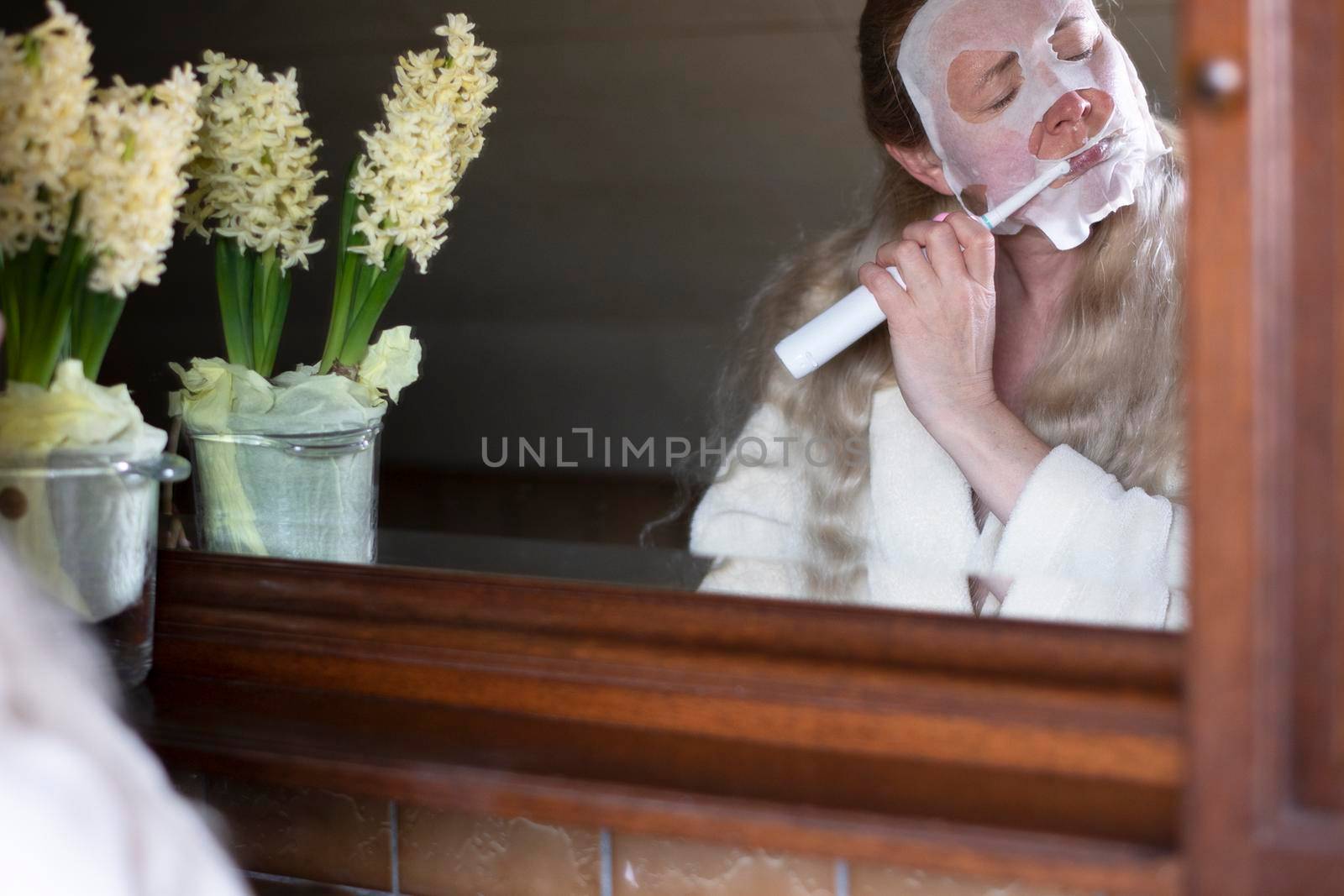 middle-aged woman in a refreshing mask is busy with morning hygiene, brushing her teeth with an electric toothbrush. High quality photo