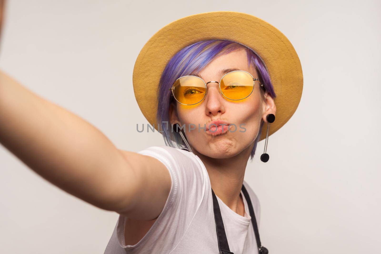 Portrait of lovely glamour hipster girl with violet hair in hat and sunglasses taking selfie, looking at camera POV, point of view of photo, sending air kiss. isolated on white background, studio shot
