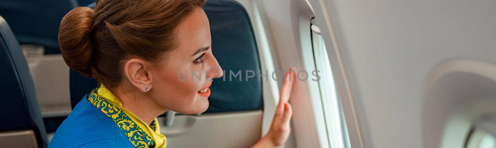 Close up of female flight attendant enjoying the view from aircraft window and smiling while standing near passenger seat