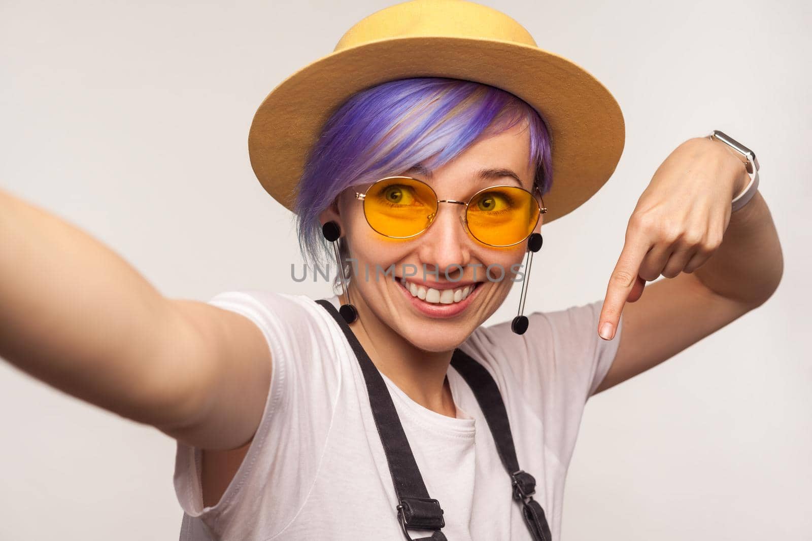 Portrait of attractive glamour hipster girl with violet hair in hat and sunglasses taking selfie or making video call, looking at camera POV, point of view of photo, pointing finger down. studio shot