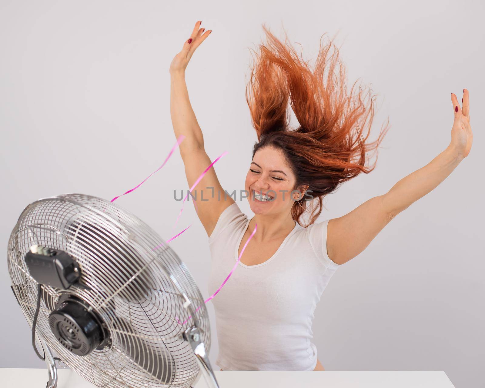 Joyful caucasian woman enjoying the wind blowing from an electric fan on a white background