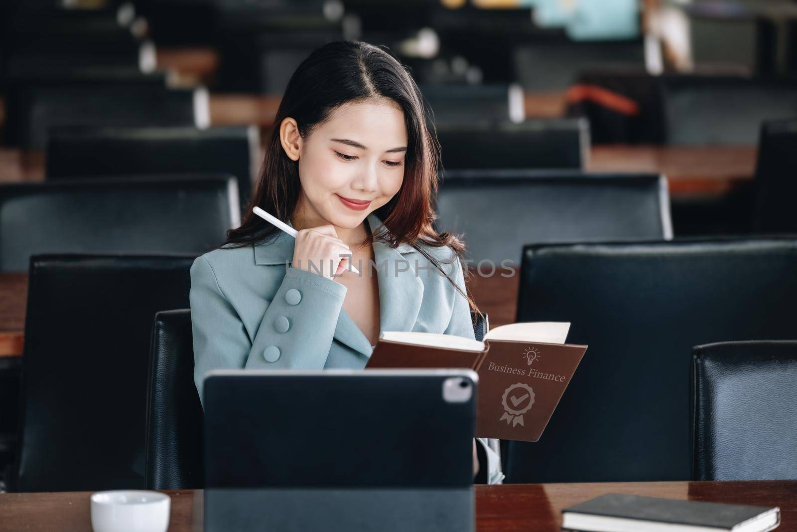 Asia woman entrepreneur showing a smiling face while reading a book developing financial and investing strategies and operating a computer tablet working on a wooden table. by Manastrong