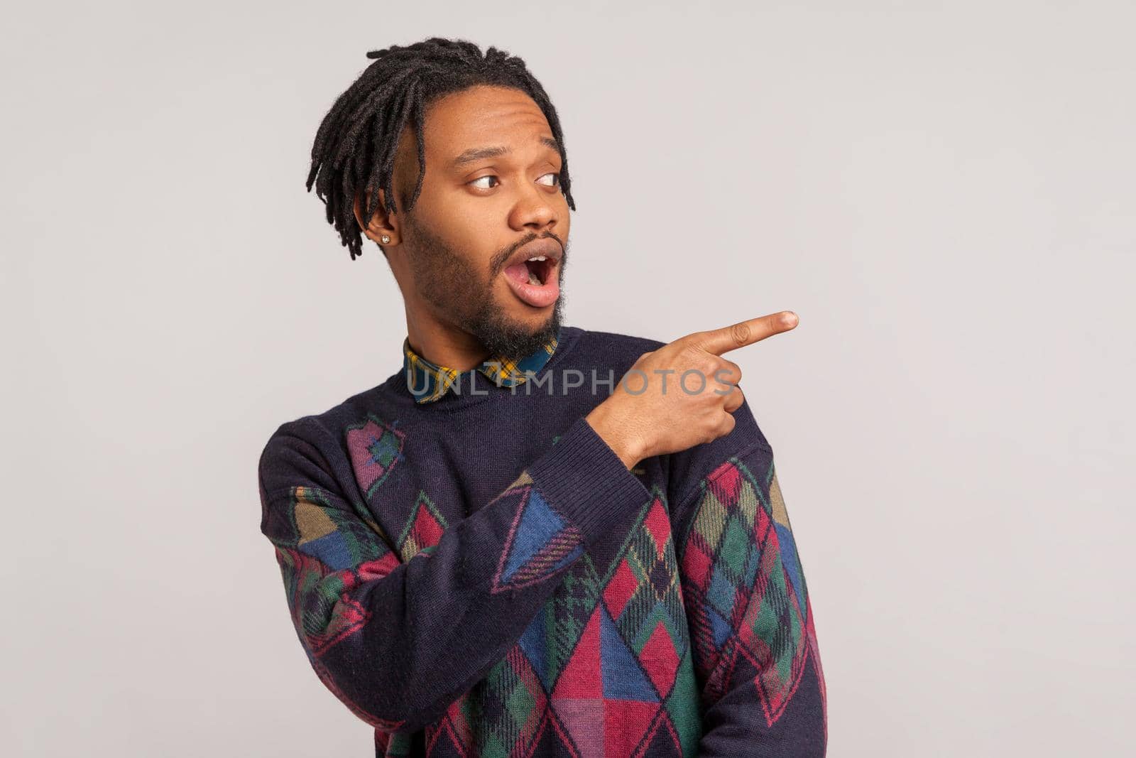 Extremely surprised african guy with dreadlocks pointing finger away with big eyes and open mouth, wondered amazed with advertisement, freespace. Indoor studio shot isolated on gray background