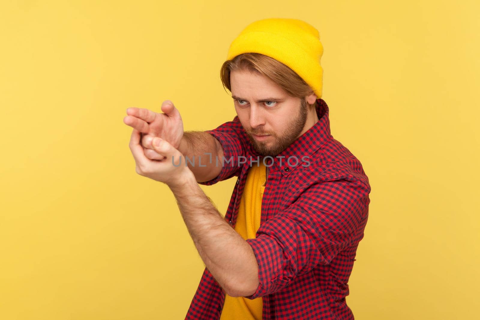 Dangerous hooligan hipster guy in beanie hat and checkered shirt pointing finger gun, threatening to kill, shooting with weapon gesture right in target. studio shot isolated on yellow background