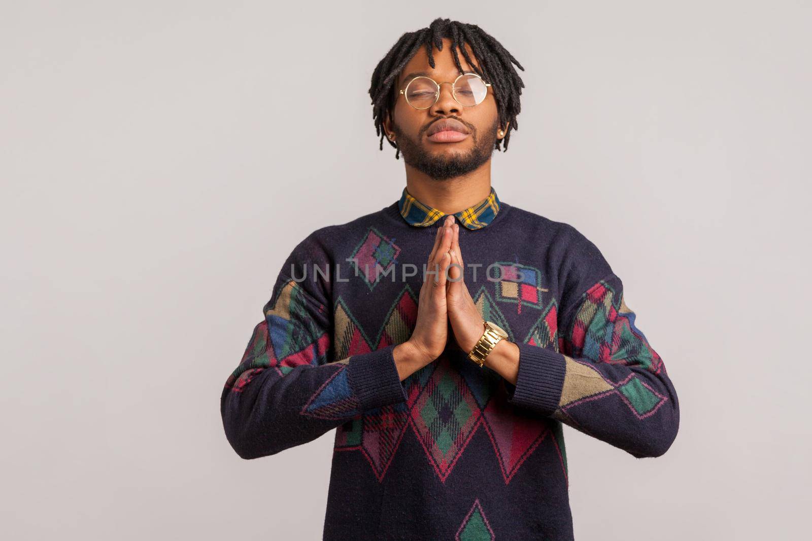 Closeup concentrated african man in glasses with dreadlocks closing eyes and pressing hands together trying to calm down, yoga practice, zen. Indoor studio shot isolated on gray background