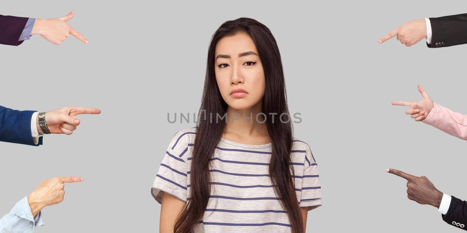 Many different people hands are pointing on guilty alone young brunette woman and blame her. she looking at camera with sad face. indoor studio shot, isolated on gray background.