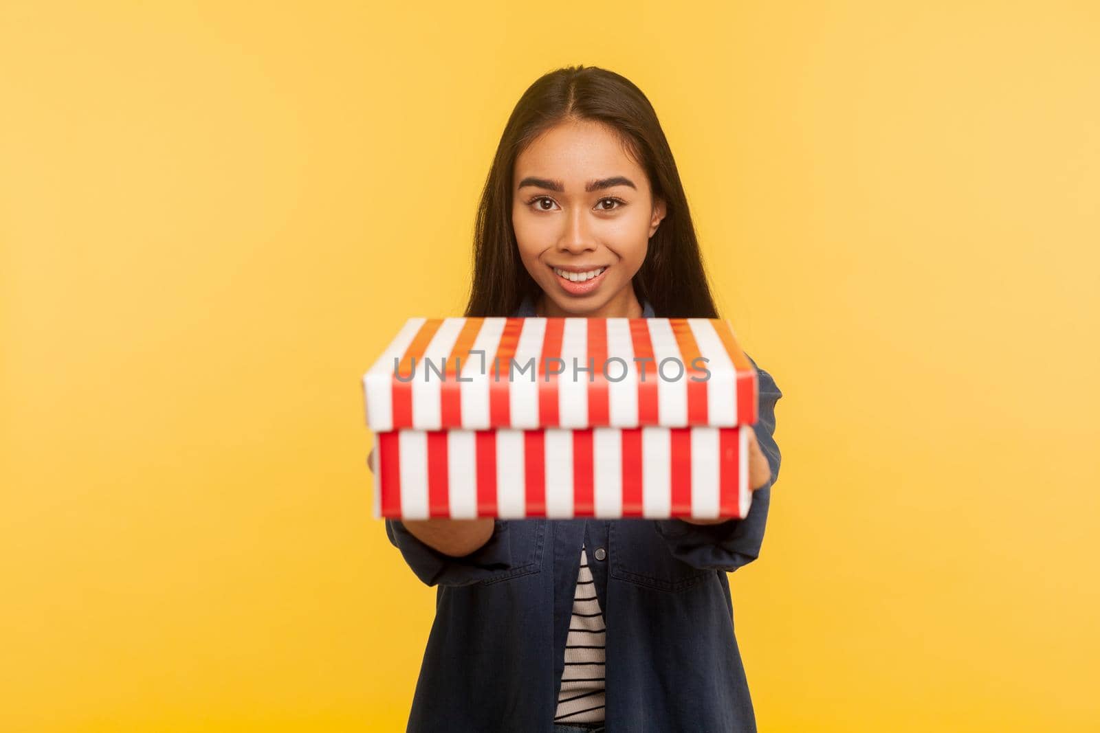 Present for you. Portrait of generous happy brunette girl with charming smile giving box to camera, sharing holiday gift, congratulating on birthday. indoor studio shot isolated on yellow background