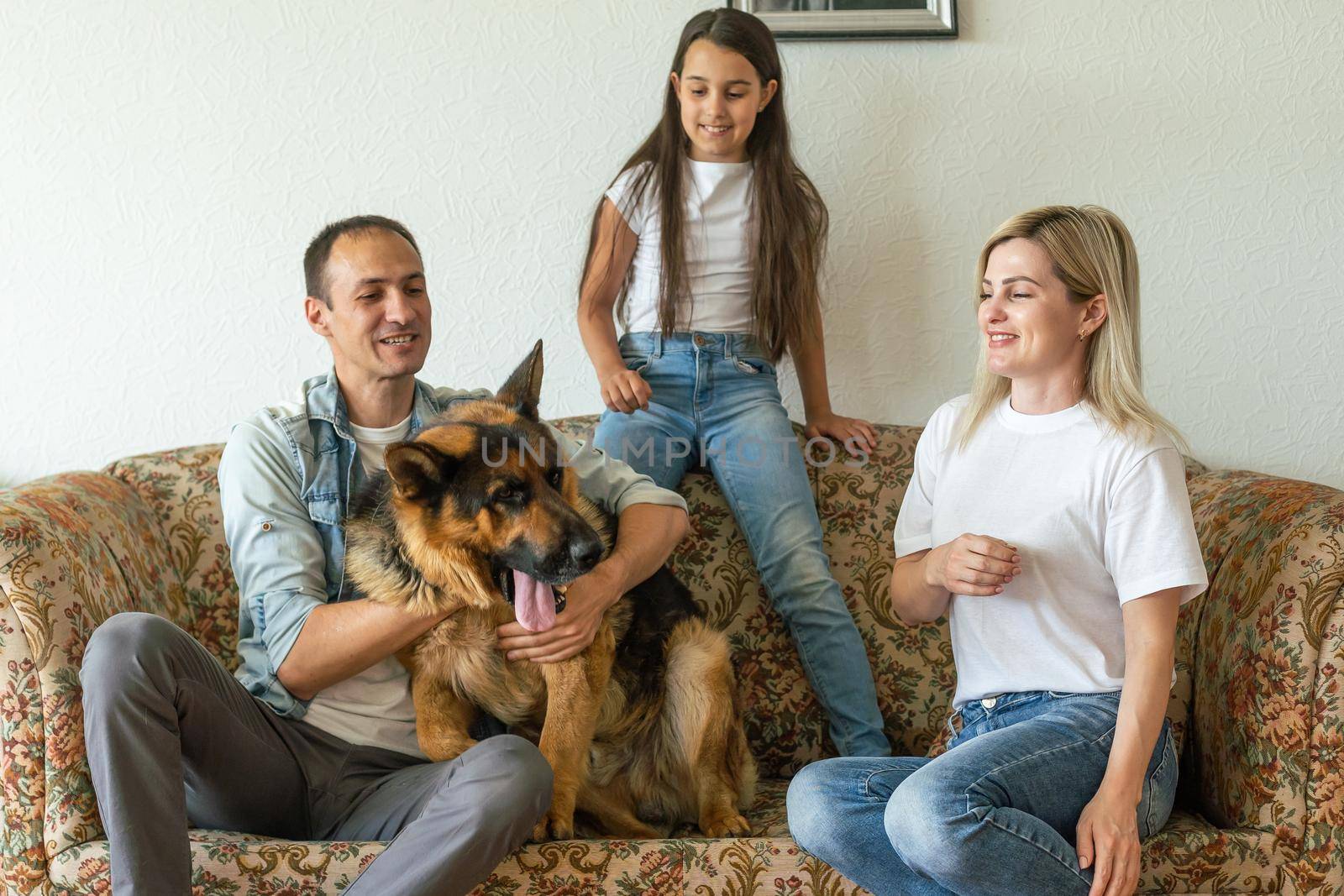 Beautiful young parents, their cute little daughter looking at camera and smiling, sitting with their cute dog on sofa at home.