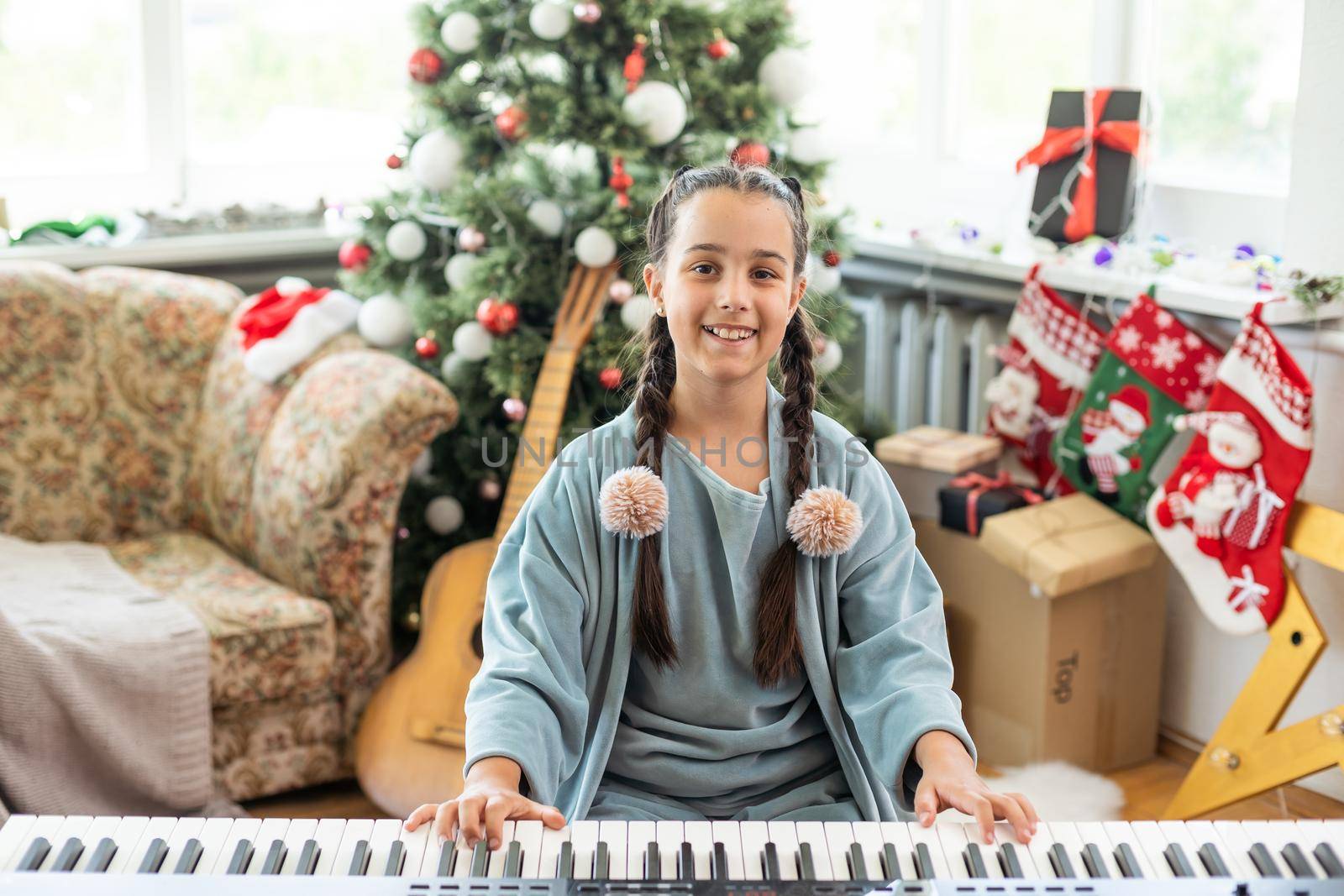 A cute little girl sitting near a white piano and a Christmas tree. The concept of the New Year, family holidays