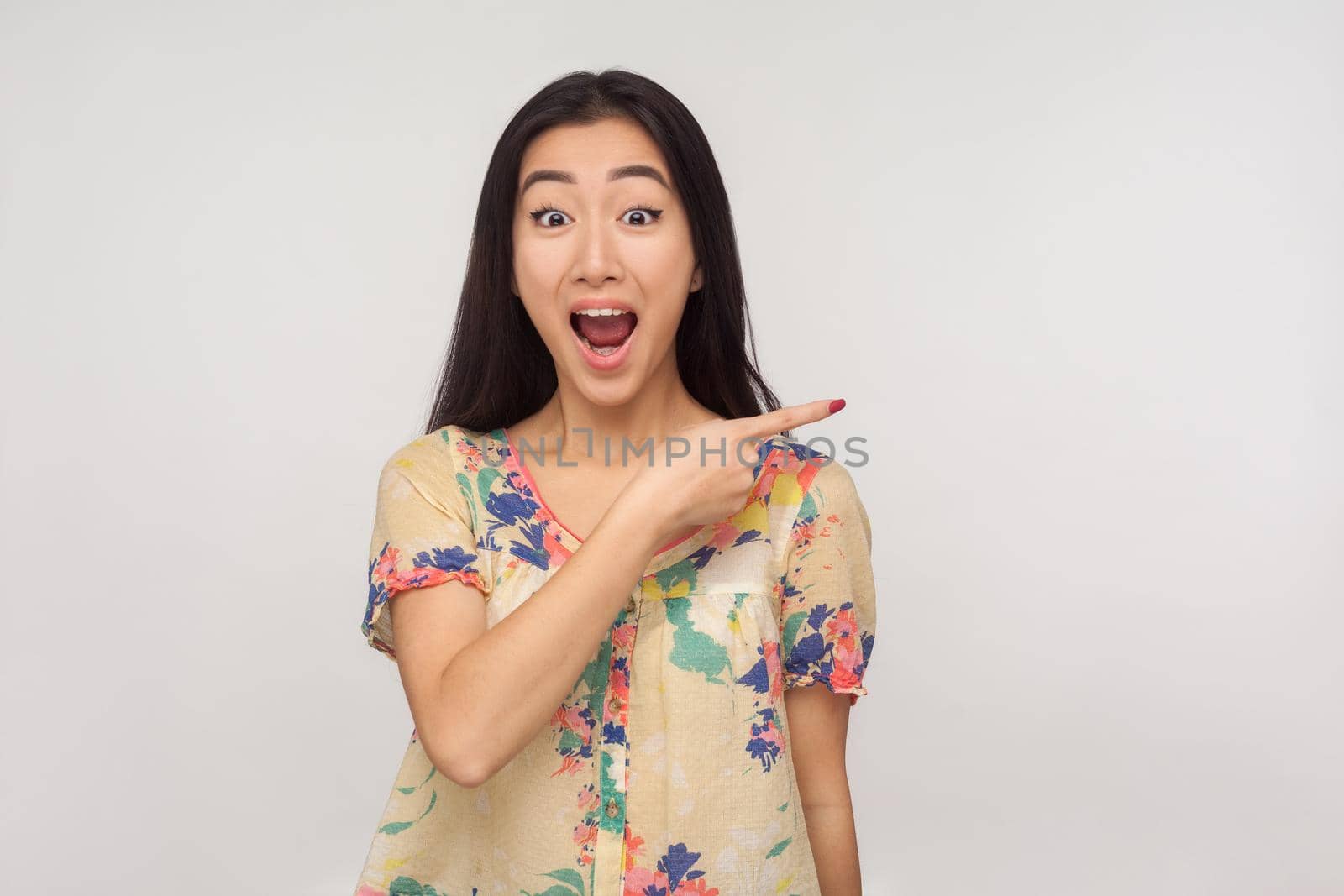 Look, awesome advertise here, Fascinating asian girl with brunette hair in summer blouse pointing aside and looking with shocked expression, showing copy space, empty place for commercial. studio shot