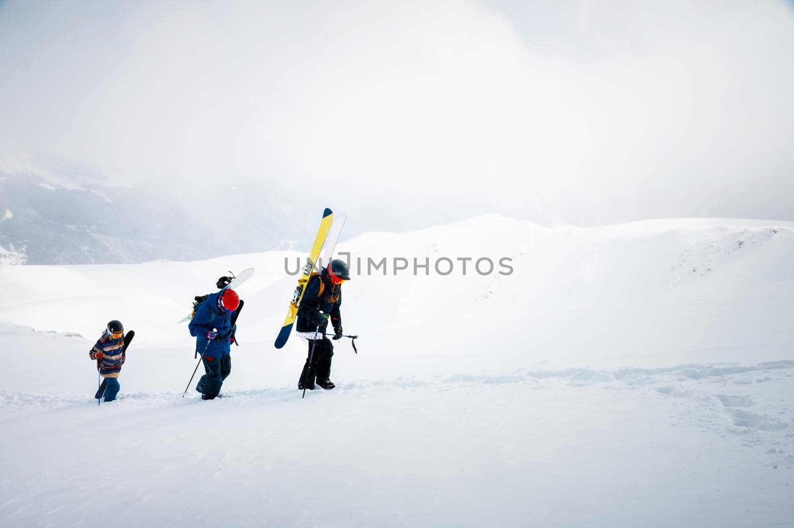 Three friends snowboarders skiers go uphill with a snowboard and skis in their hands for backcountry or freeride against the backdrop of the snow-capped mountains of the Alpine resort. Ski tour concept with group of peoples