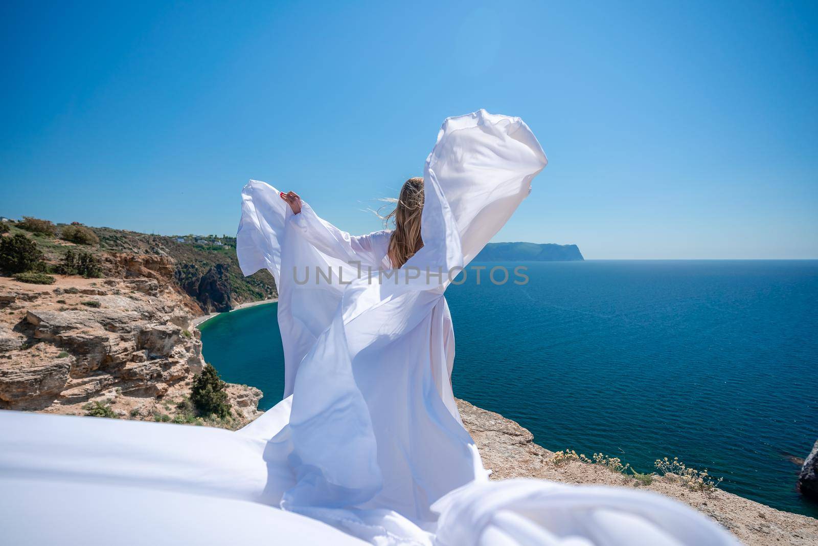 Blonde with long hair on a sunny seashore in a white flowing dress, rear view, silk fabric waving in the wind. Against the backdrop of the blue sky and mountains on the seashore