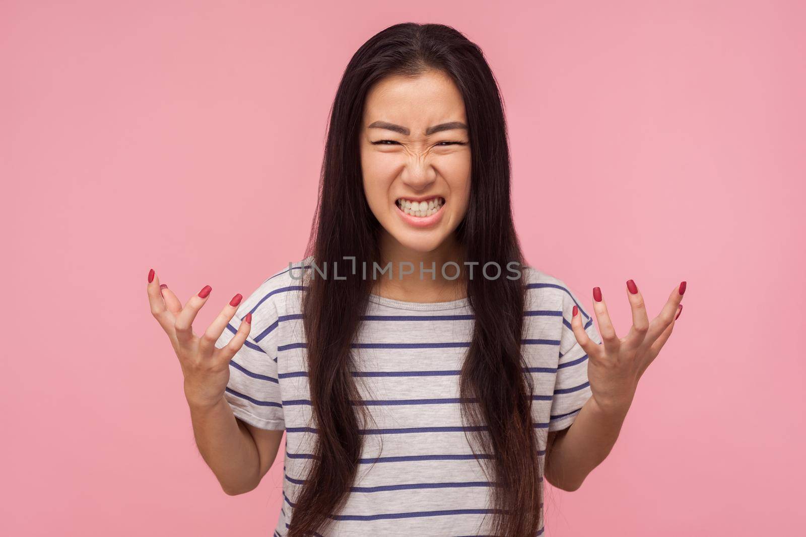 Portrait of enraged furious girl with long hair in striped t-shirt standing with clenched teeth and raised hands expressing wild anger, feeling crazy. indoor studio shot isolated on pink background