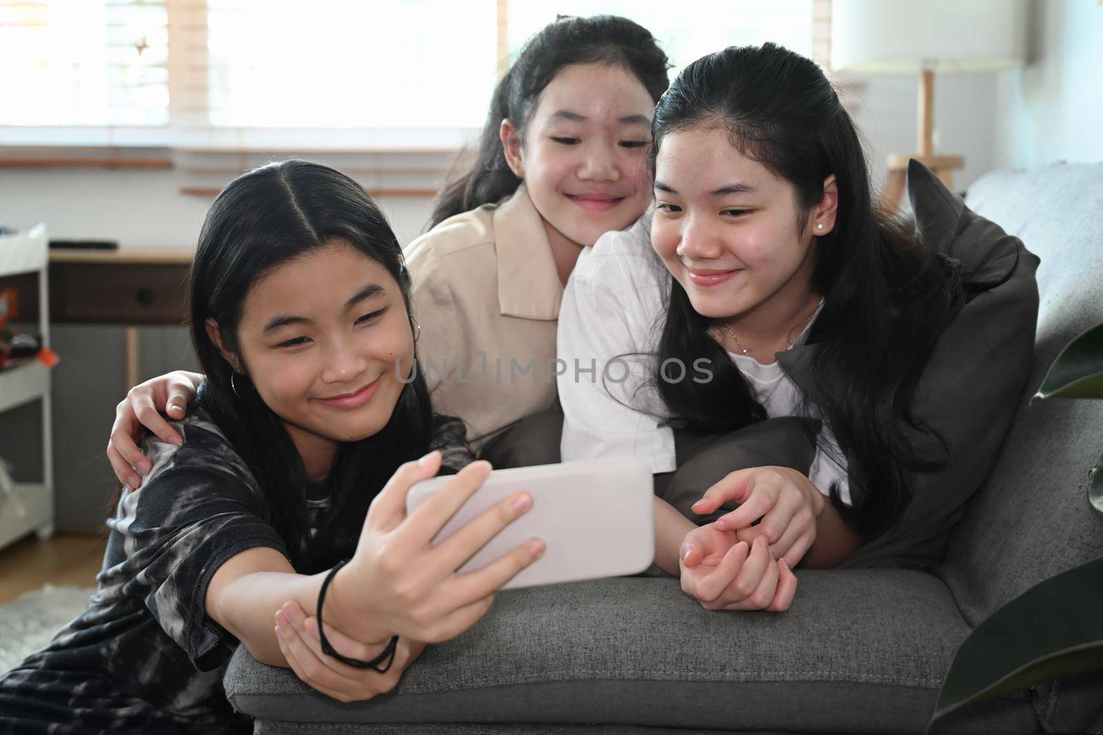 Cheerful Asian girls taking selfie with smart smartphone while sitting together in living room. by prathanchorruangsak
