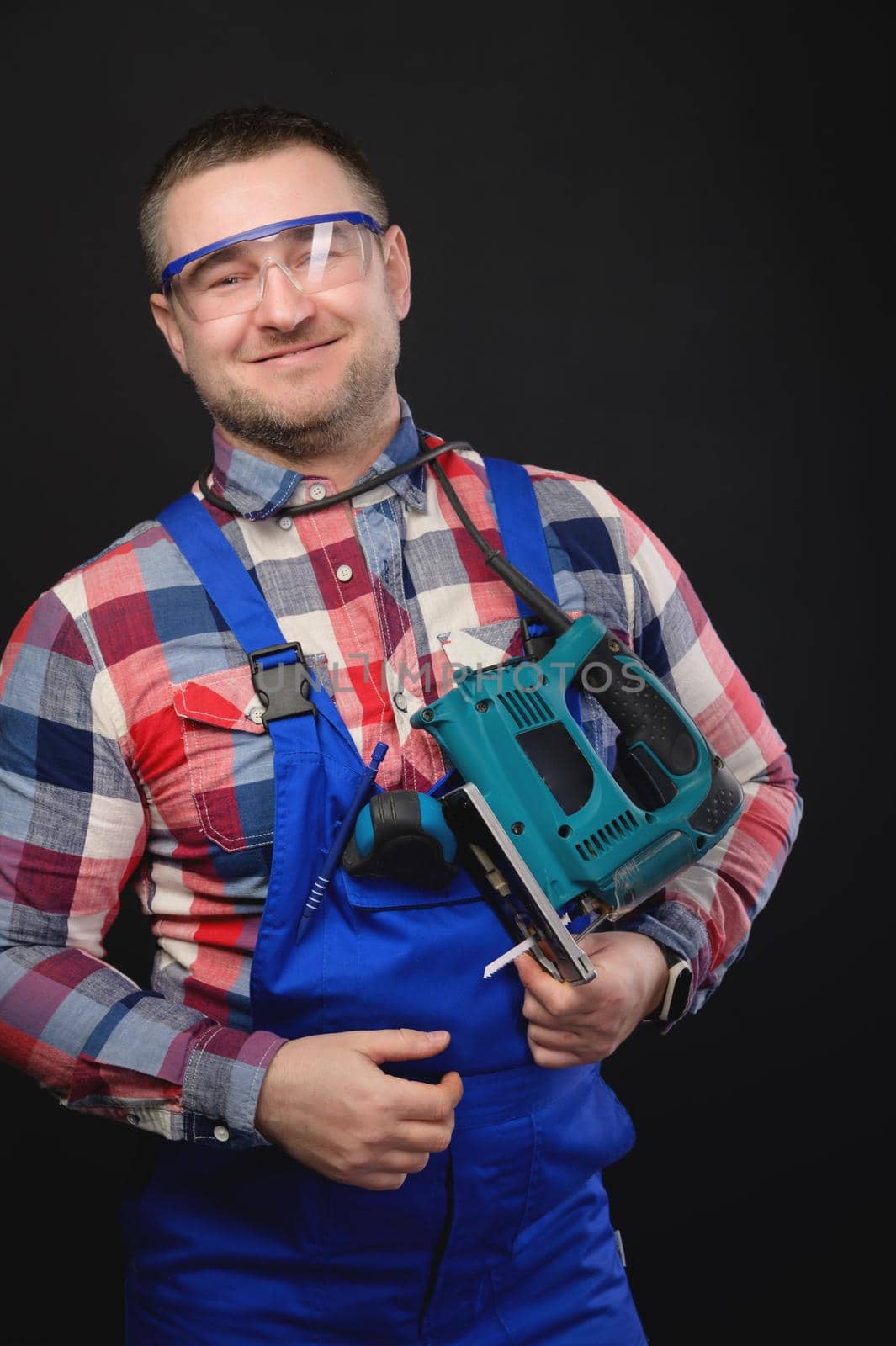 Portrait of a caucasian male repairman in a work uniform with an electric jigsaw around his neck. Studio portrait of an artisan businessman.