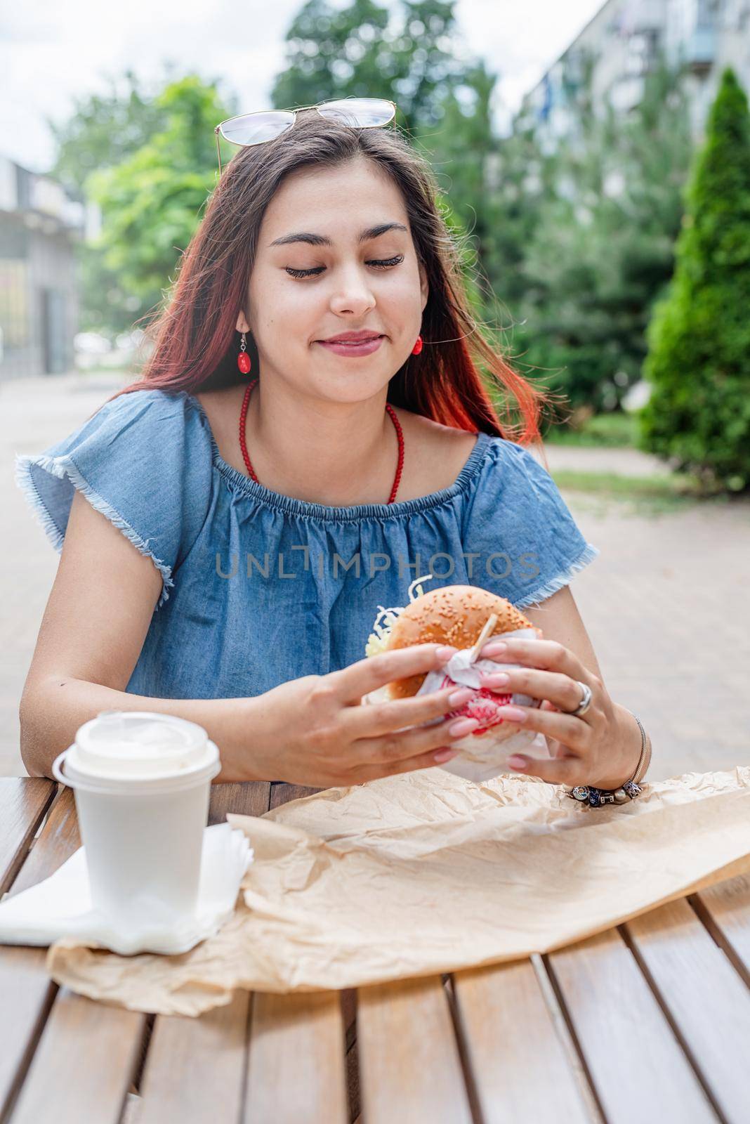 Summer vacation, street food eating. Charming hungry stylish woman, enjoying eating a burger outdoors, dressed in jeans shirt, wearing sunglasses