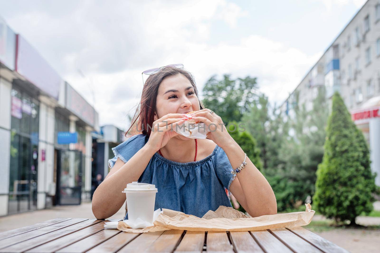 Summer vacation, street food eating. Charming hungry stylish woman, enjoying eating a burger outdoors, dressed in jeans shirt, wearing sunglasses