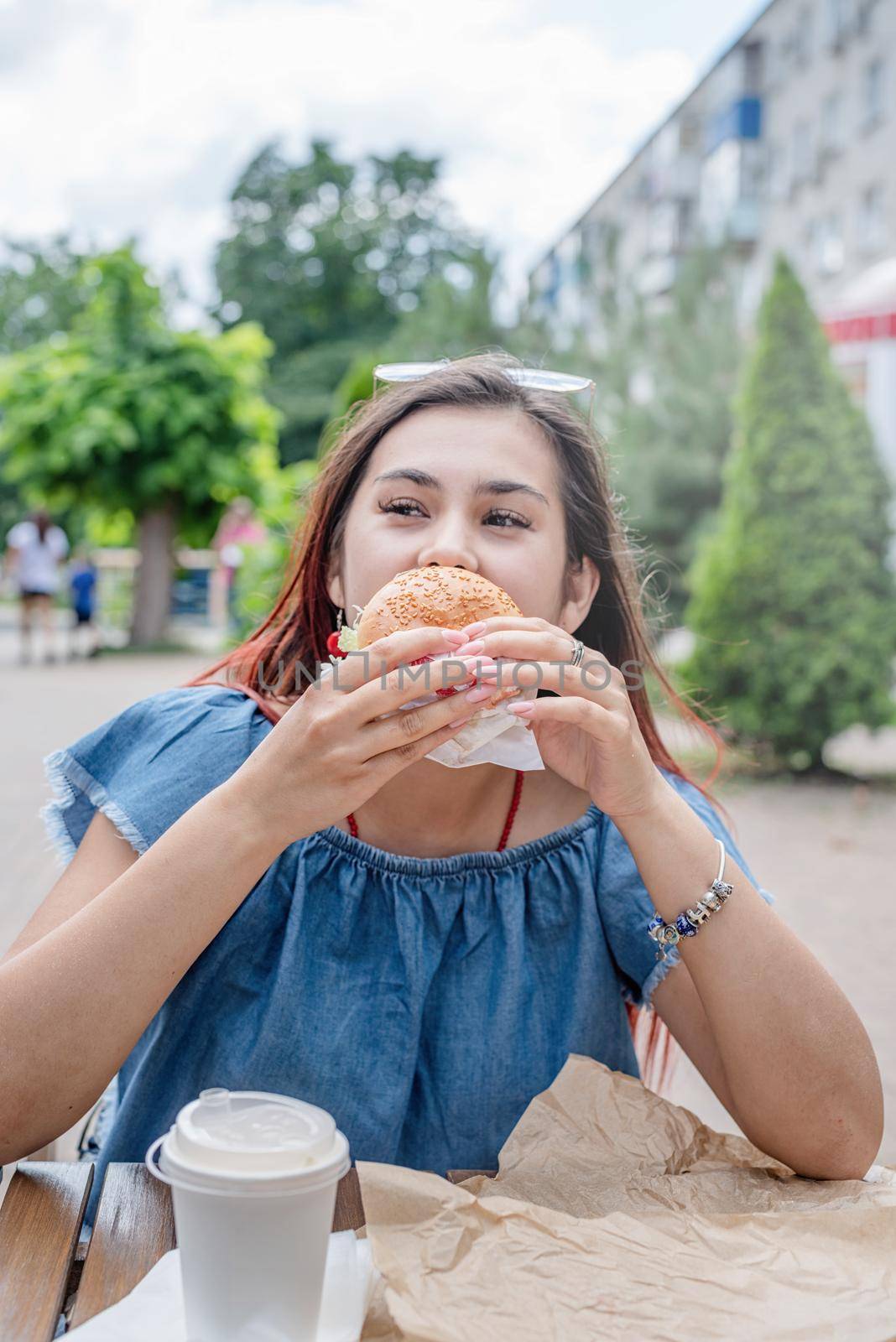 Summer vacation, street food eating. Charming hungry stylish woman, enjoying eating a burger outdoors, dressed in jeans shirt, wearing sunglasses