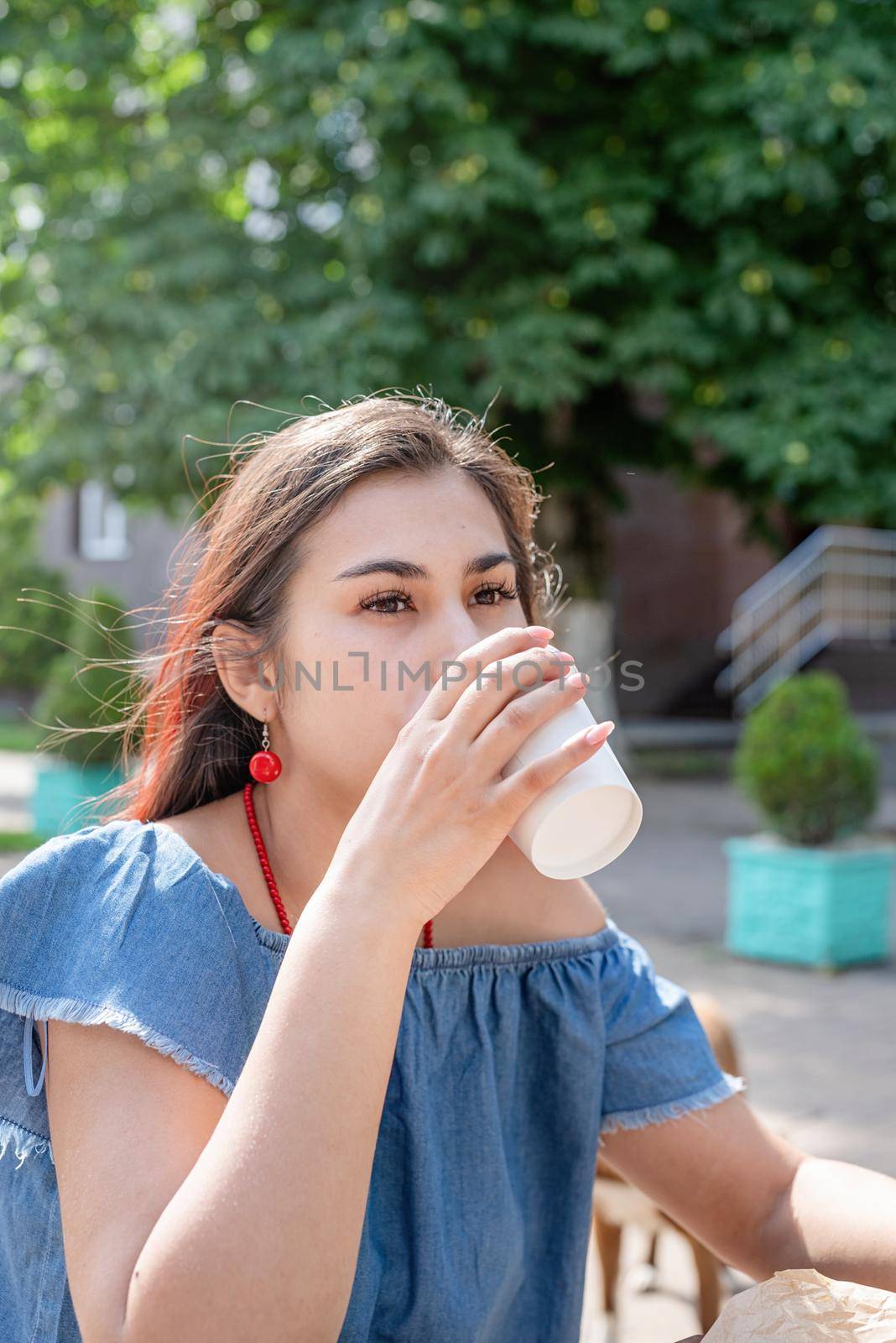 Summer vacation, street food eating. Charming hungry stylish woman, enjoying drinking coffee outdoors, dressed in jeans shirt, wearing sunglasses