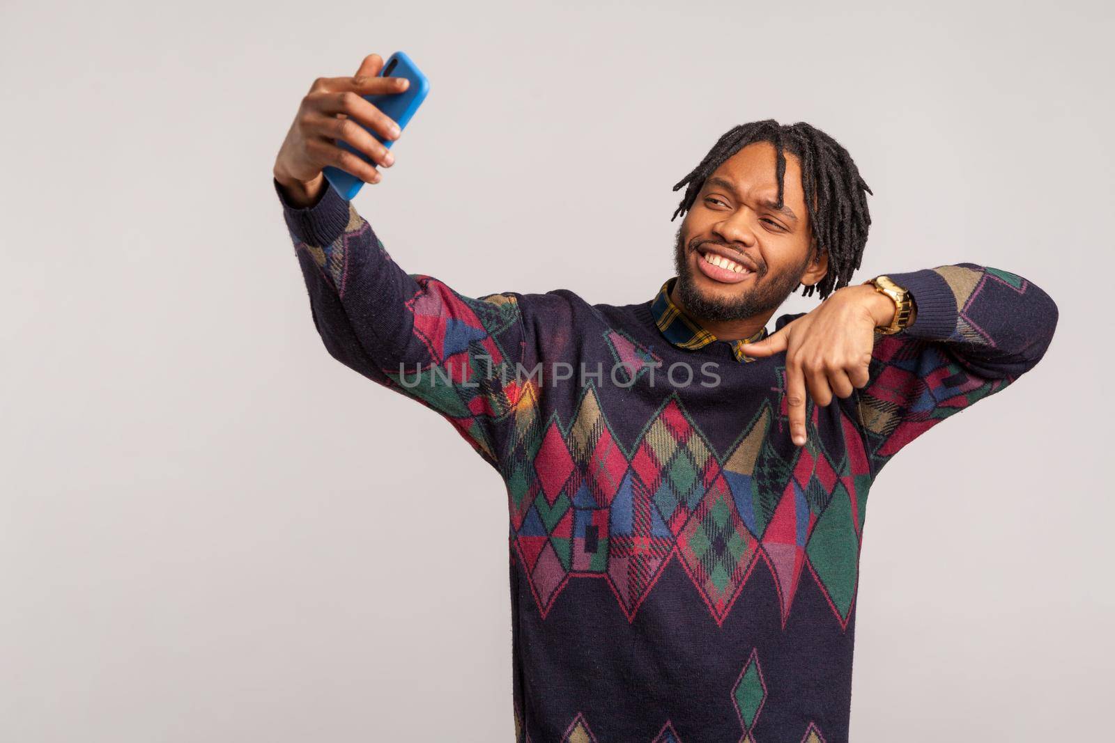 Handsome self confident african male blogger with dreadlocks showing subscribe gesture looking at camera of smartphone with toothy smile. Indoor studio shot isolated on gray background