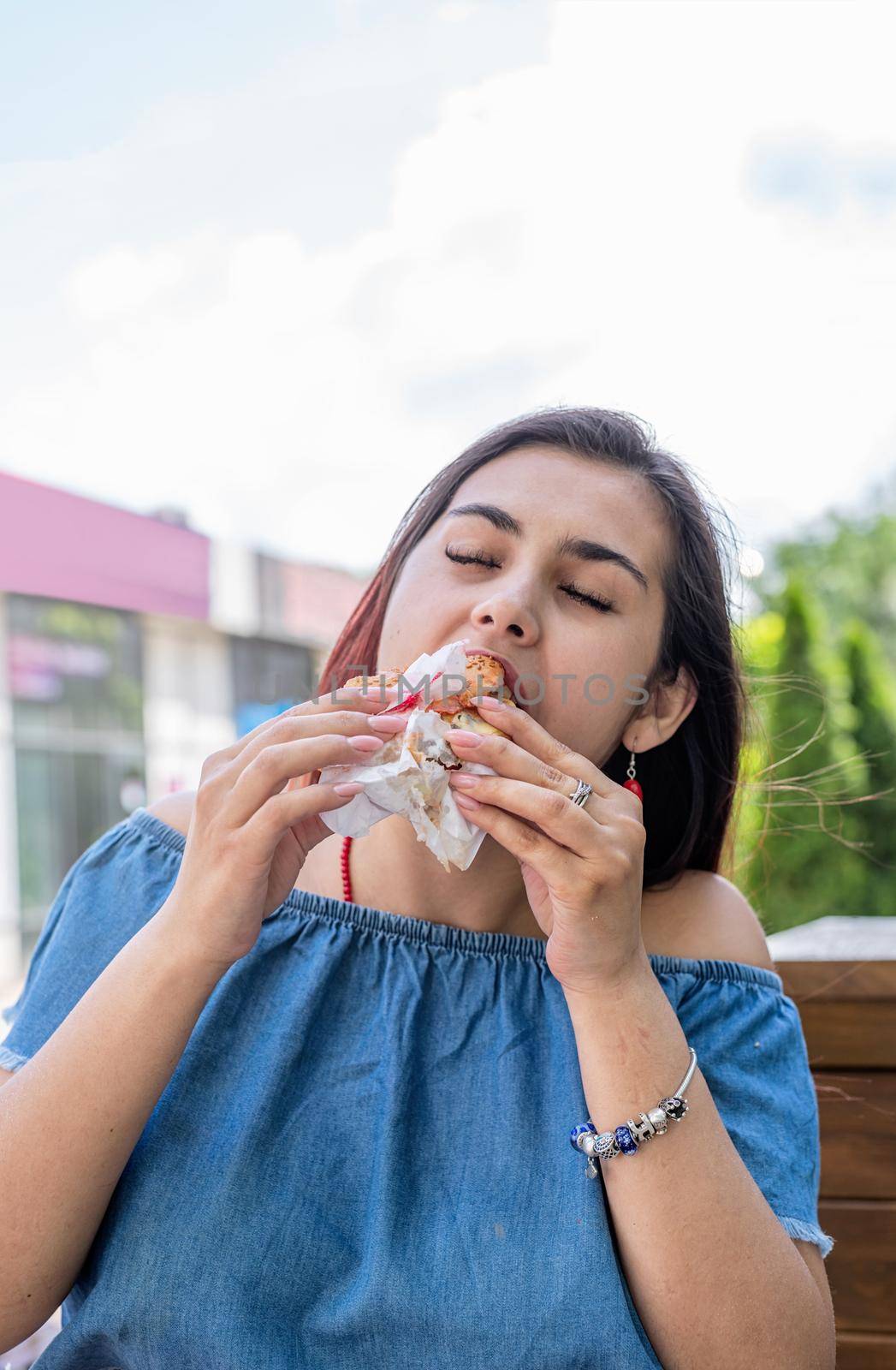 Summer vacation, street food eating. Charming hungry stylish woman, enjoying eating a burger outdoors, dressed in jeans shirt, wearing sunglasses