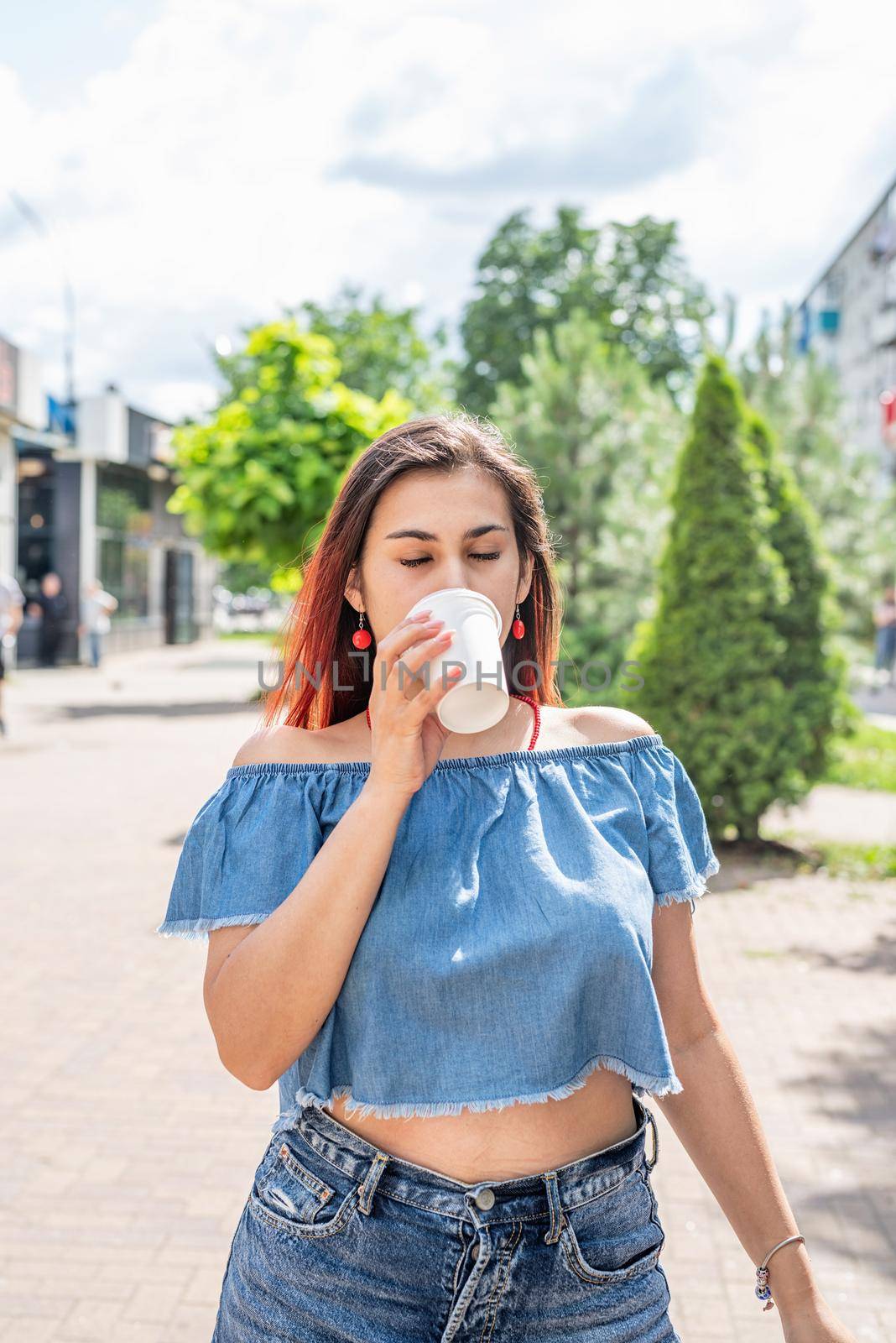 Summer vacation, street food eating. Charming hungry stylish woman, enjoying drinking coffee outdoors, dressed in jeans shirt, wearing sunglasses