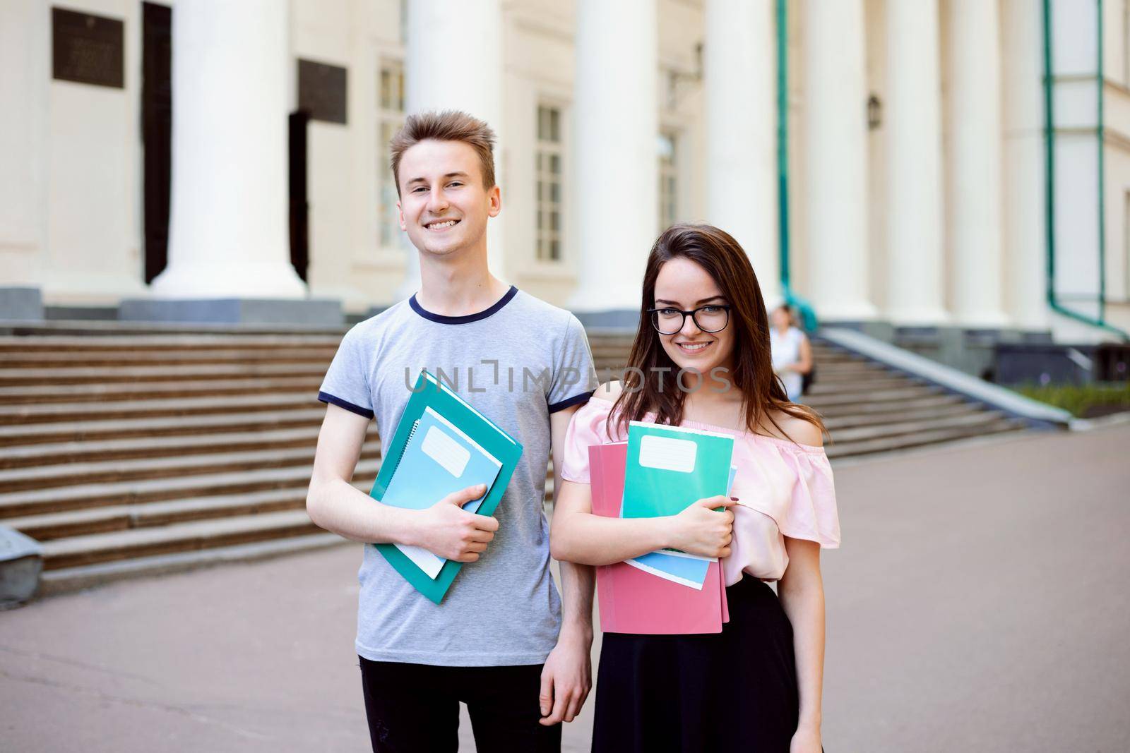 Two exemplary students in front of old beautiful conventional university with high columns posing with learning materials dressed in casual clothes looks happy and encouraged to study