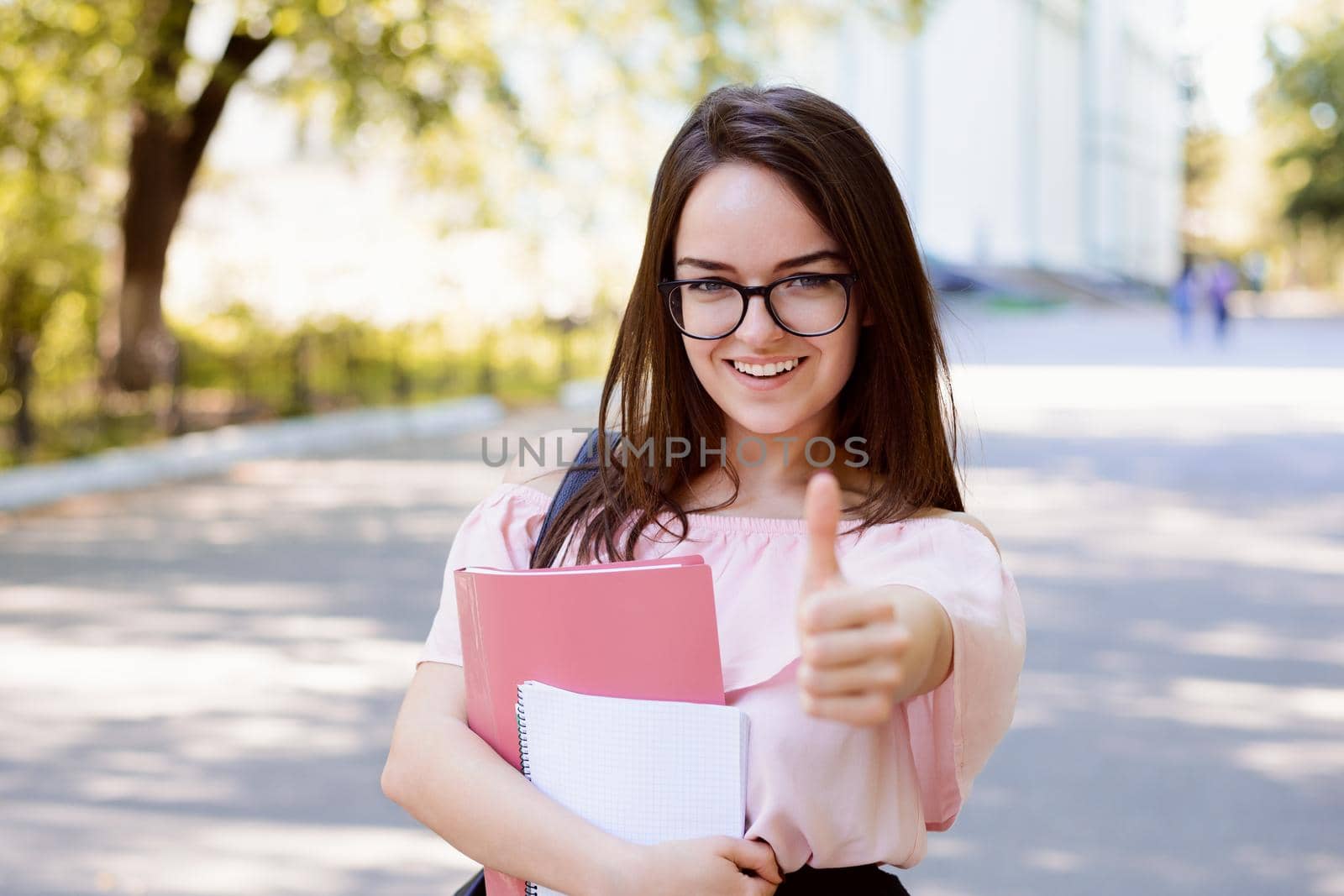 Student showing thumbs up standing against old conventional university with books and notes, wearing spectacles, blouse and skirt expressing happiness
