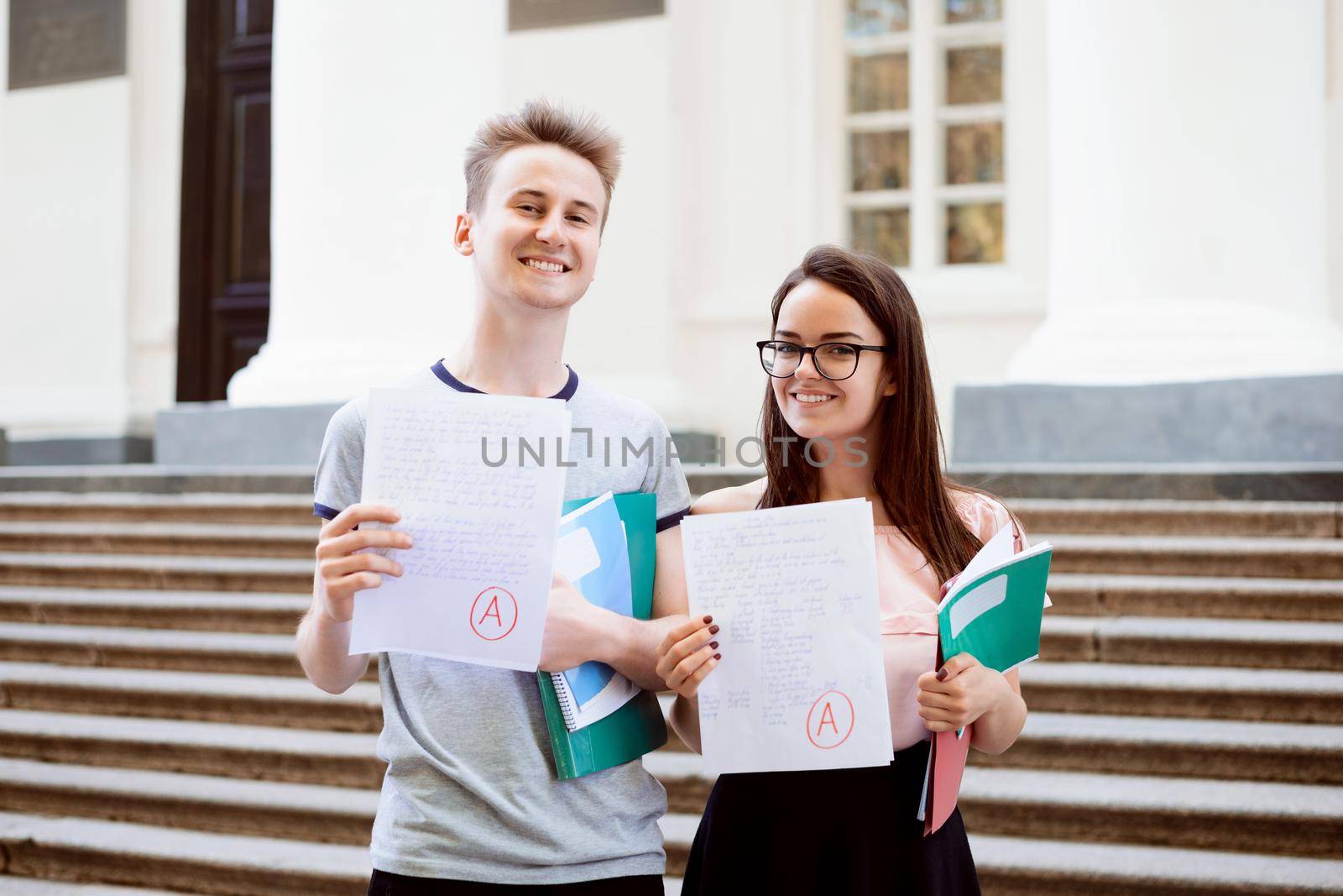 Male and female teenagers standing in front of university showing excellent results of entrance exam, happy to become students of a popular university and study hard