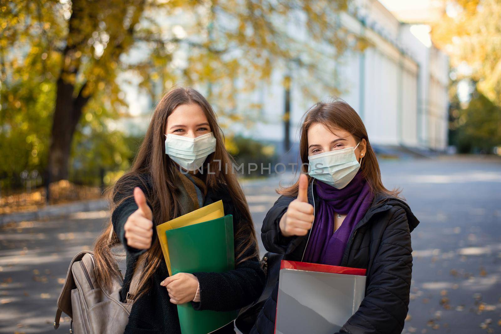 Two female students in protective medical masks showing thumbs up to the camera standing in university campus by VitaliiPetrushenko