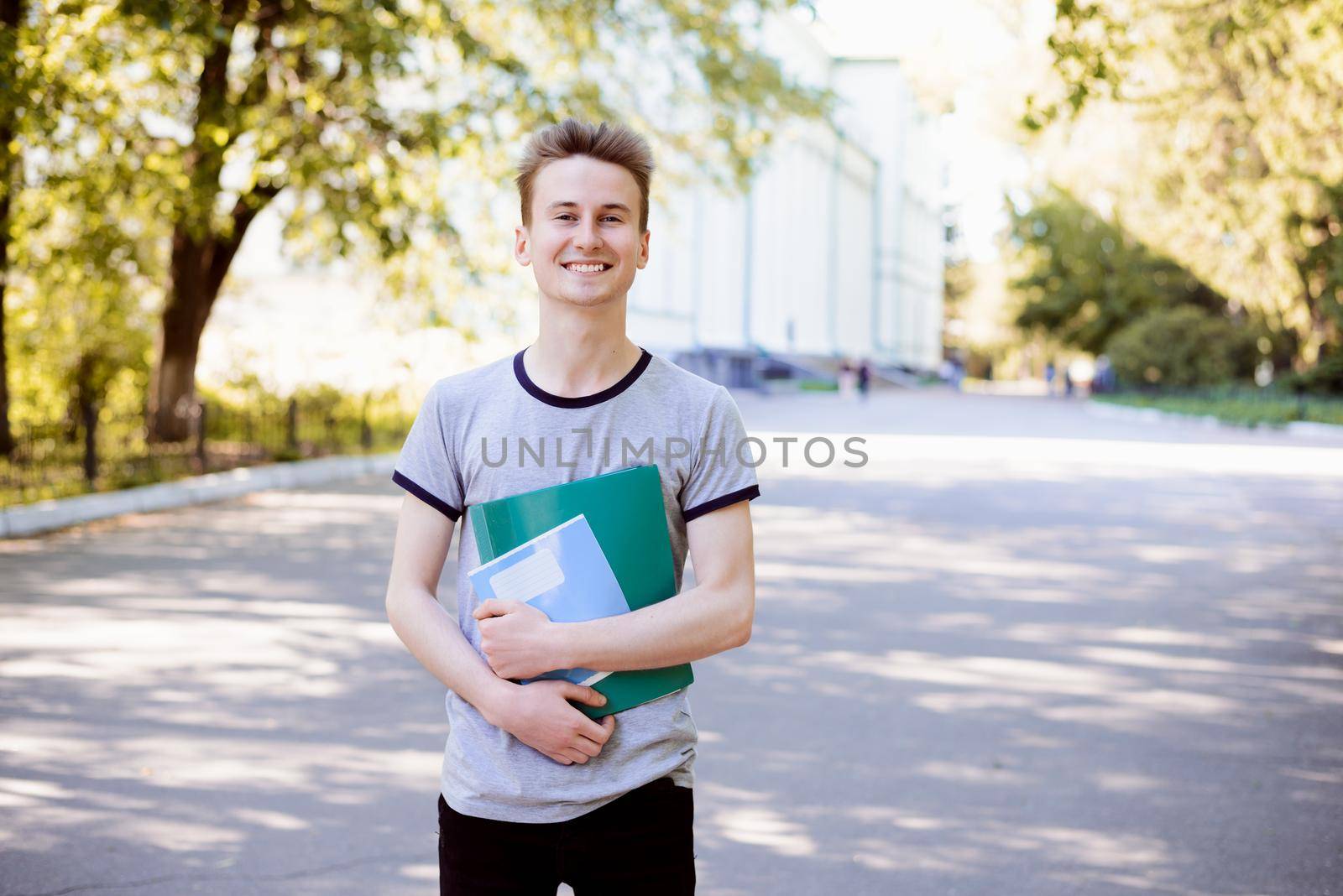 Cheerful young student with folder and exercise books enjoying walk in the park near university in the evening after classes