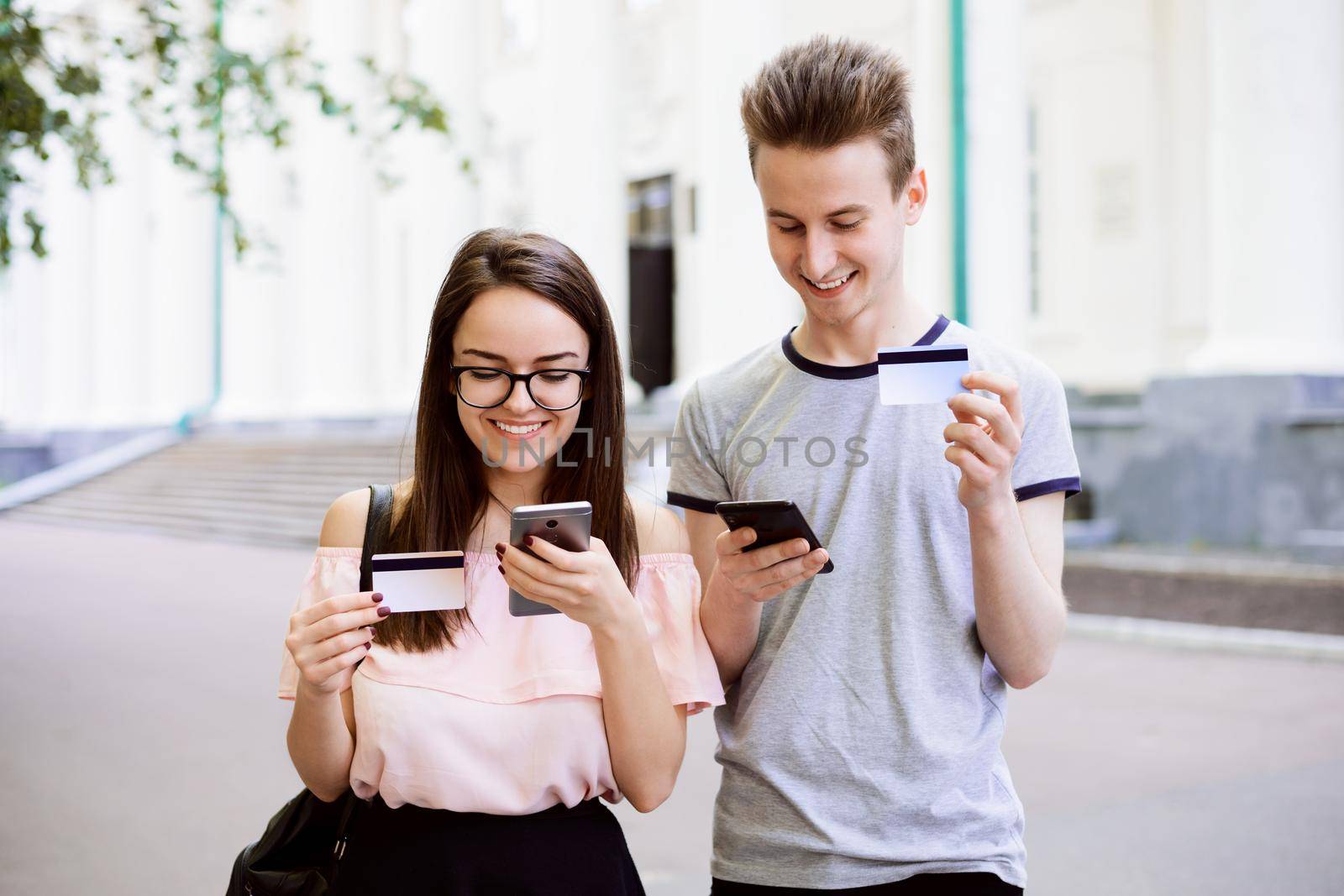Attractive male and female standing in front of conventional university with credit cards and mobile phones in hands, purchasing things in the web